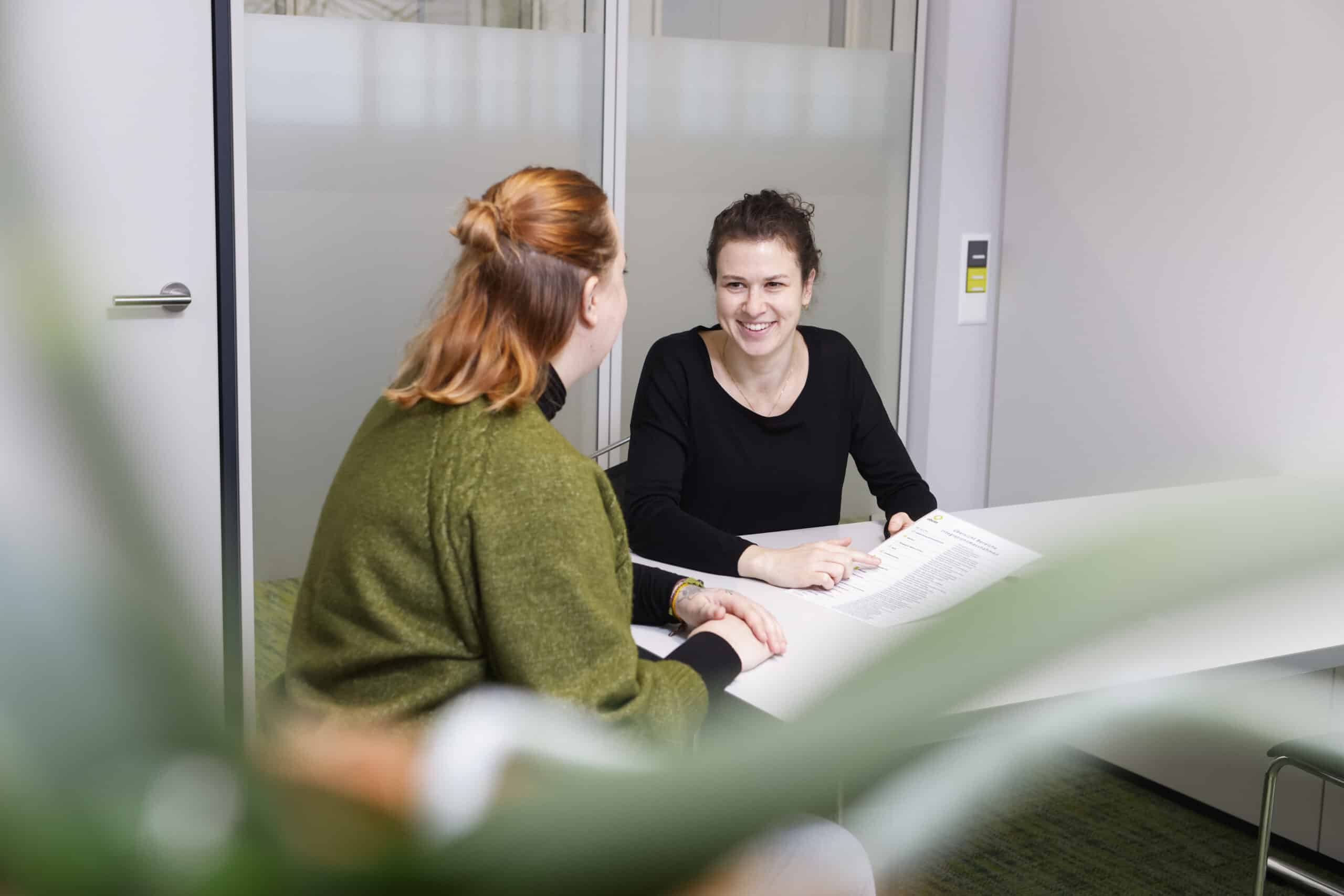 Zwei Frauen sitzen sich an einem weißen Tisch in einem hellen Büro gegenüber, lächeln und unterhalten sich. Eine hält ein Dokument in der Hand. Im Vordergrund ist unscharf eine Blattpflanze zu erkennen.