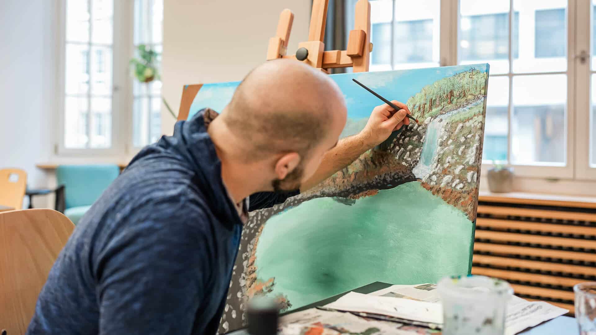 Eine Person mit Glatze malt eine Landschaft mit Wasser, Bäumen und einem Wasserfall auf eine Leinwand, die auf einer Staffelei in einem gut beleuchteten Raum steht.
