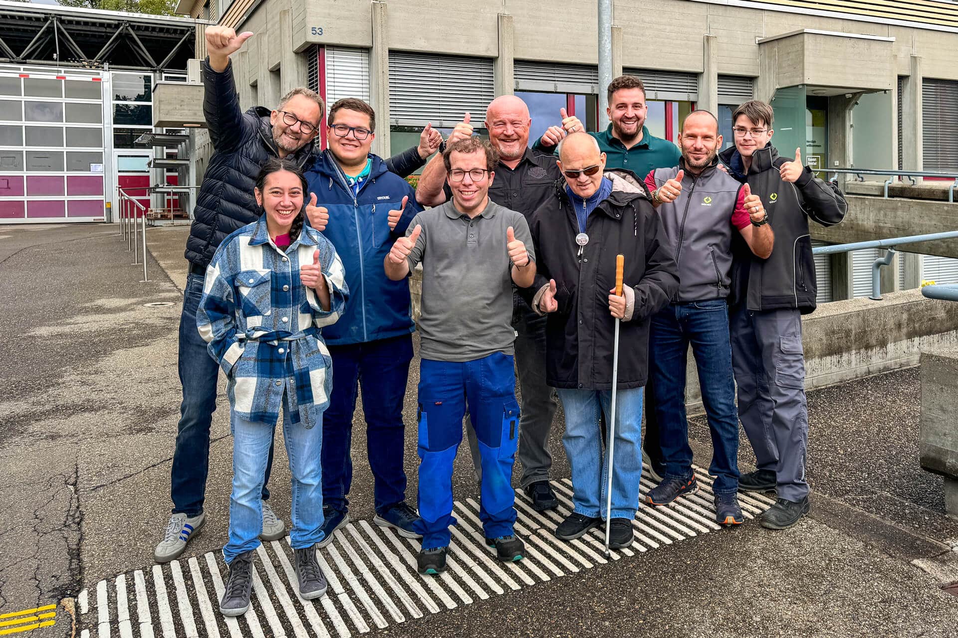 Eine Gruppe von zehn Personen steht draußen auf einem Bürgersteig, lächelt und zeigt die Daumen nach oben. Ein Mann im Vordergrund benutzt einen weißen Gehstock und trägt eine Sonnenbrille. Die Gruppe wirkt glücklich und fröhlich, mit Gebäuden im Hintergrund.