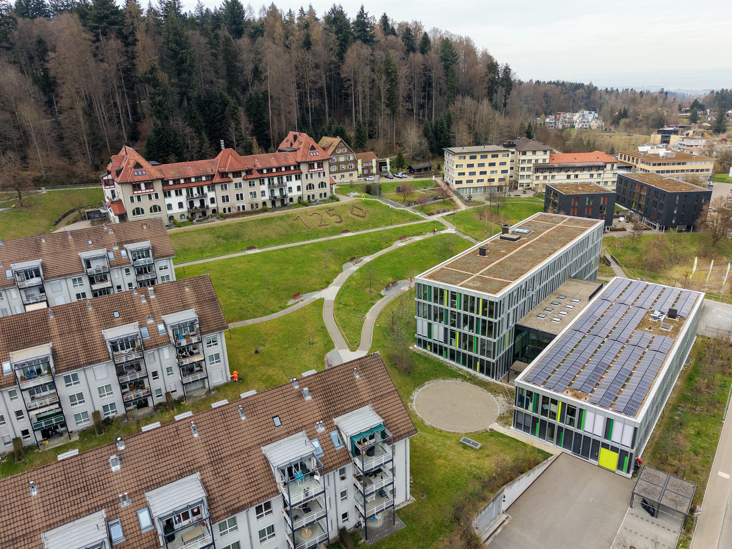 Luftaufnahme eines Campus mit modernen und älteren Gebäuden, grünen Rasenflächen und Wegen, umgeben von Bäumen. Auf den Dächern befinden sich Solarzellen, und auf dem Rasen ist "1250" zu sehen.