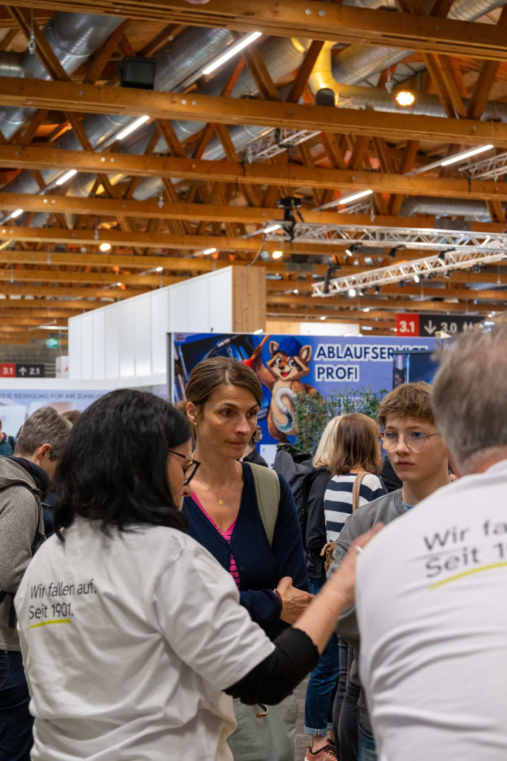Eine Gruppe von Personen unterhält sich bei einer Veranstaltung in einem Innenraum mit hölzernen Deckenbalken darüber. Zwei Personen tragen weiße Hemden mit deutscher Aufschrift. Im Hintergrund sind ein blauer Stand mit einem Bärenmaskottchen und weitere Teilnehmer zu sehen.