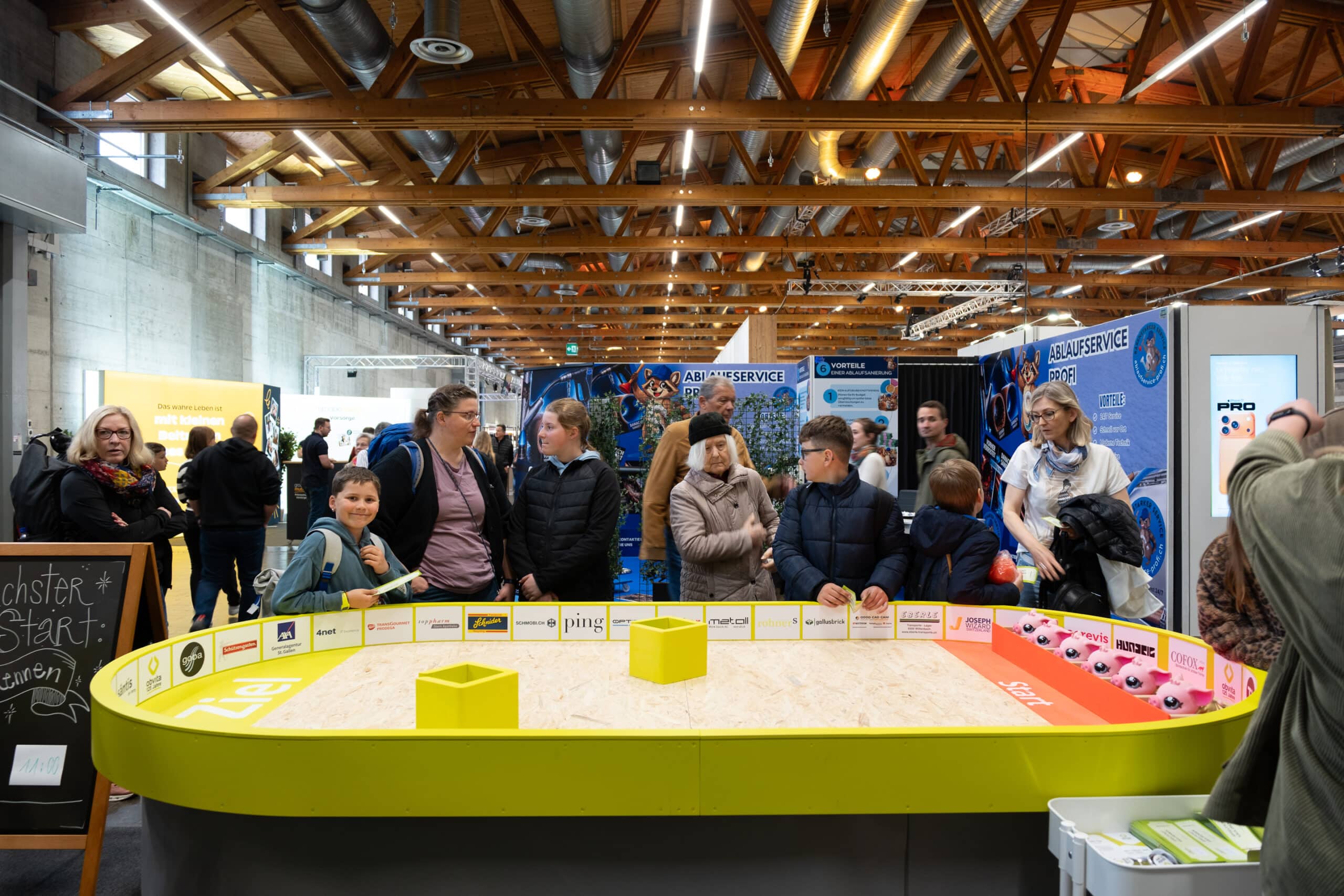 Eine Gruppe von Menschen, darunter auch Kinder, steht um einen farbenfrohen Ausstellungstisch auf einer belebten Indoor-Veranstaltung oder Ausstellung mit verschiedenen Ständen, Bannern und Informationsschildern im Hintergrund.