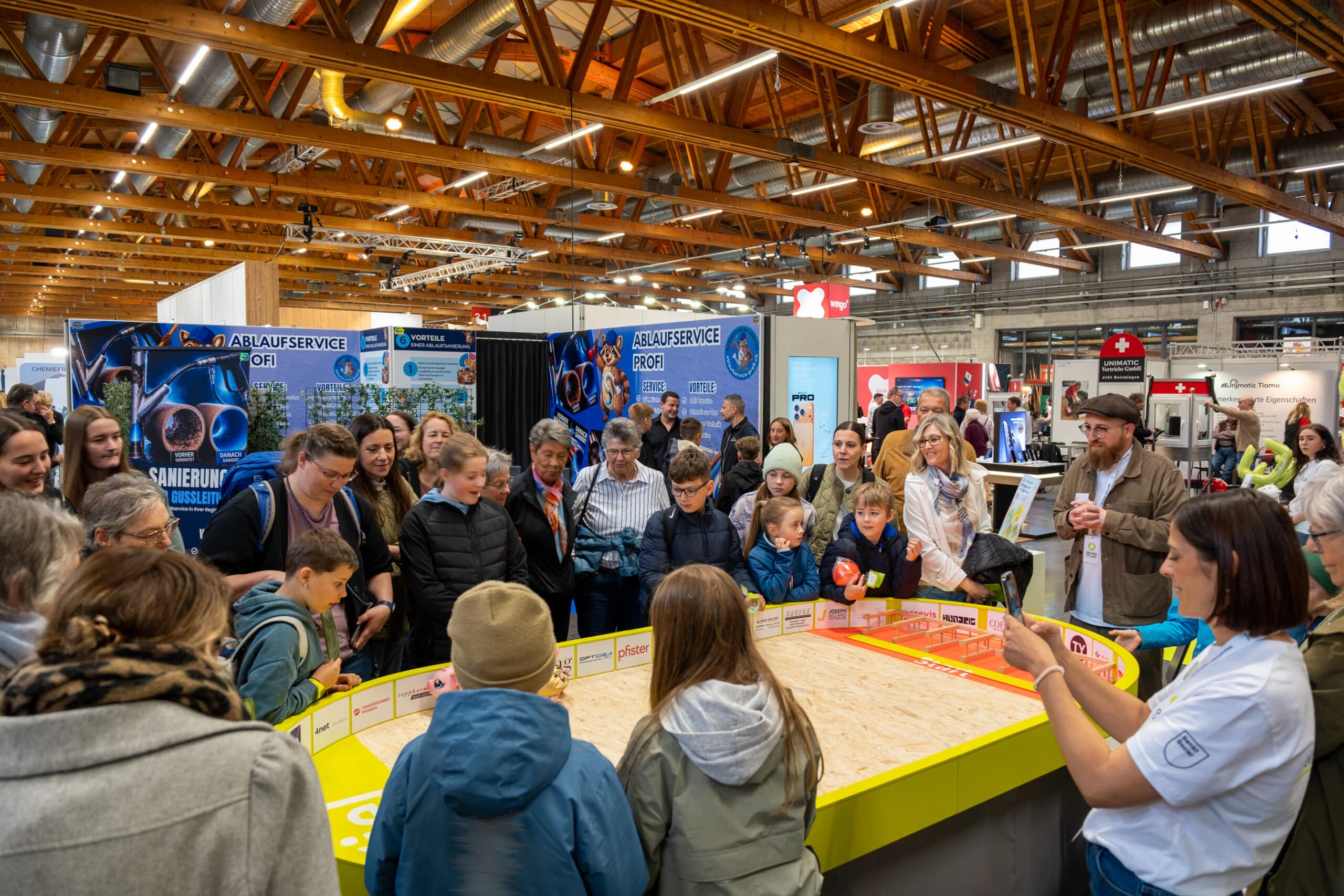 Eine Menschenmenge, darunter Kinder und Erwachsene, versammelt sich um eine Tischrobotervorführung in einer belebten Wissenschafts- oder Technologieausstellung mit Informationsständen im Hintergrund.
