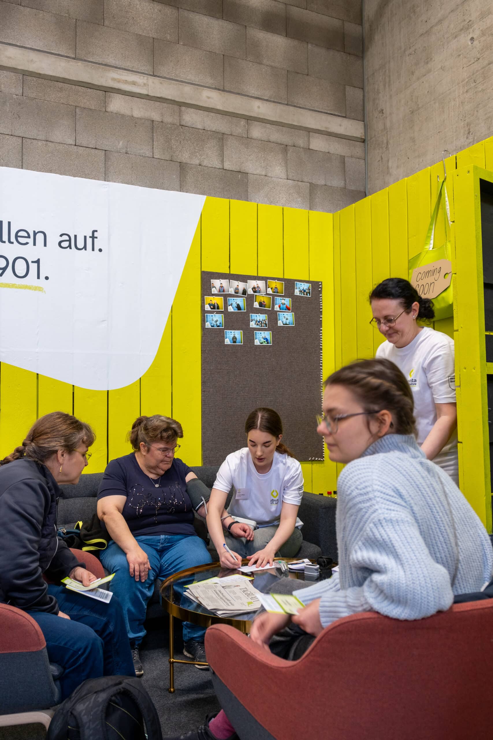 Fünf Personen sitzen in einem hellen Raum mit gelben Wänden und unterhalten sich. Zwei tragen weiße Hemden und helfen anderen; einer prüft den Blutdruck einer Frau. Auf dem Tisch liegen Papiere und medizinische Geräte. Fotos sind an die Wand gepinnt.