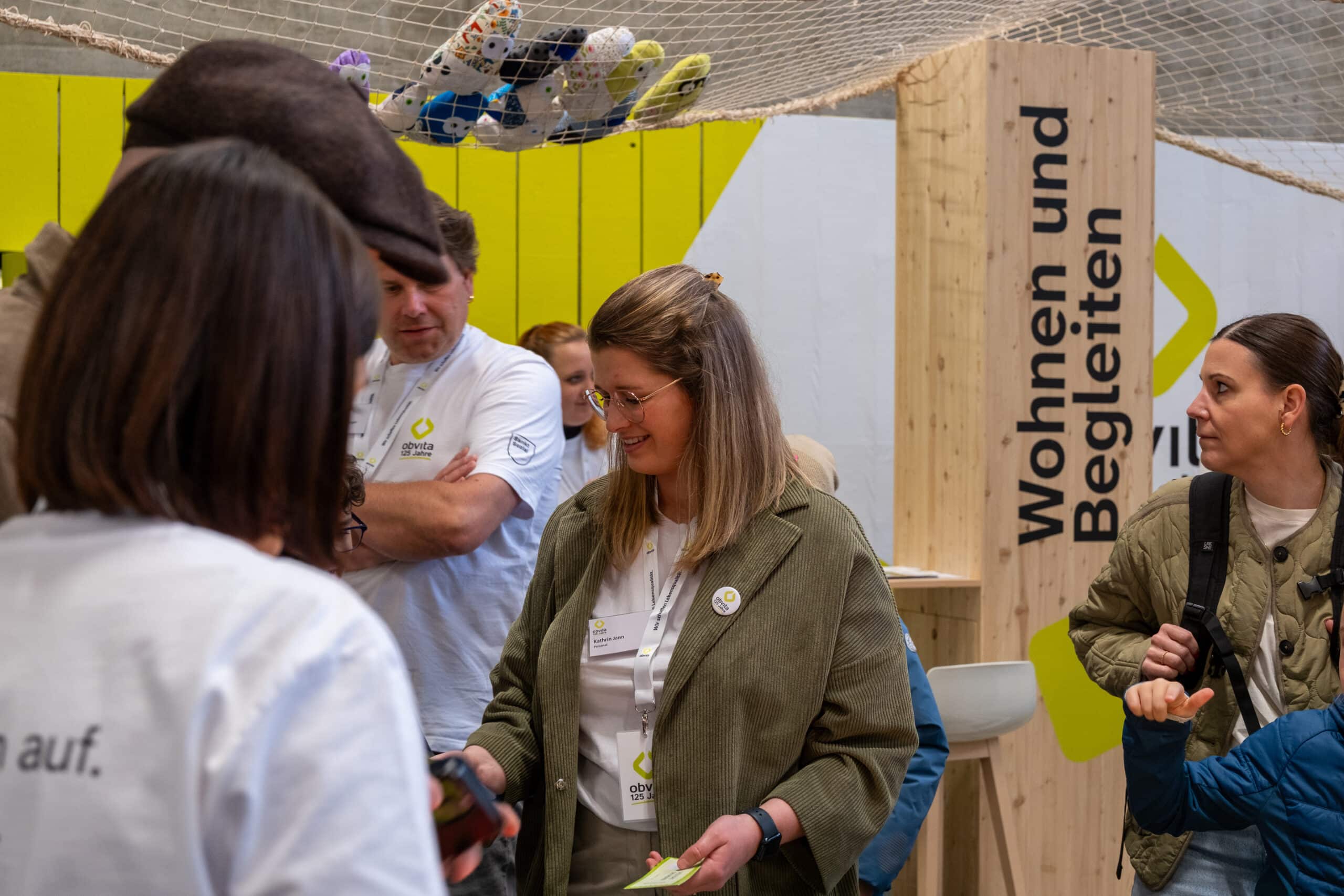 Eine Gruppe von Menschen an einem Stand für eine Indoor-Veranstaltung, die sich unterhalten und lächeln. Im Hintergrund ist ein gelbes und weißes Display mit den deutschen Worten "Wohnen und Begleiten" zu sehen. Einige Personen tragen Namensschilder und Markenshirts.