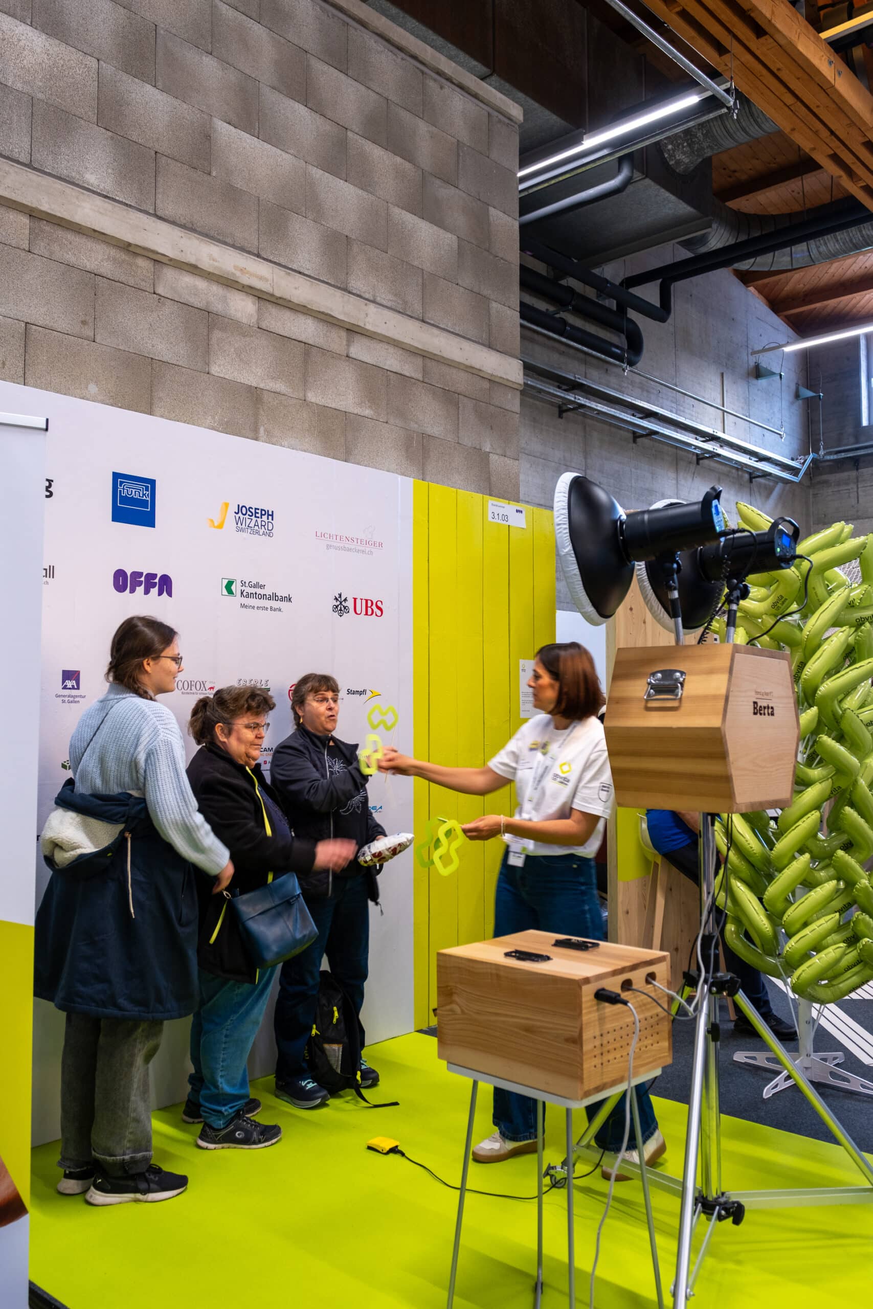 Eine Frau in einem weißen Hemd führt drei Personen, die vor einer Kulisse mit Logos stehen, bei einer Indoor-Veranstaltung das Ballonflattern vor, wobei in der Nähe ein hölzerner Lautsprecher und Ballons zu sehen sind.