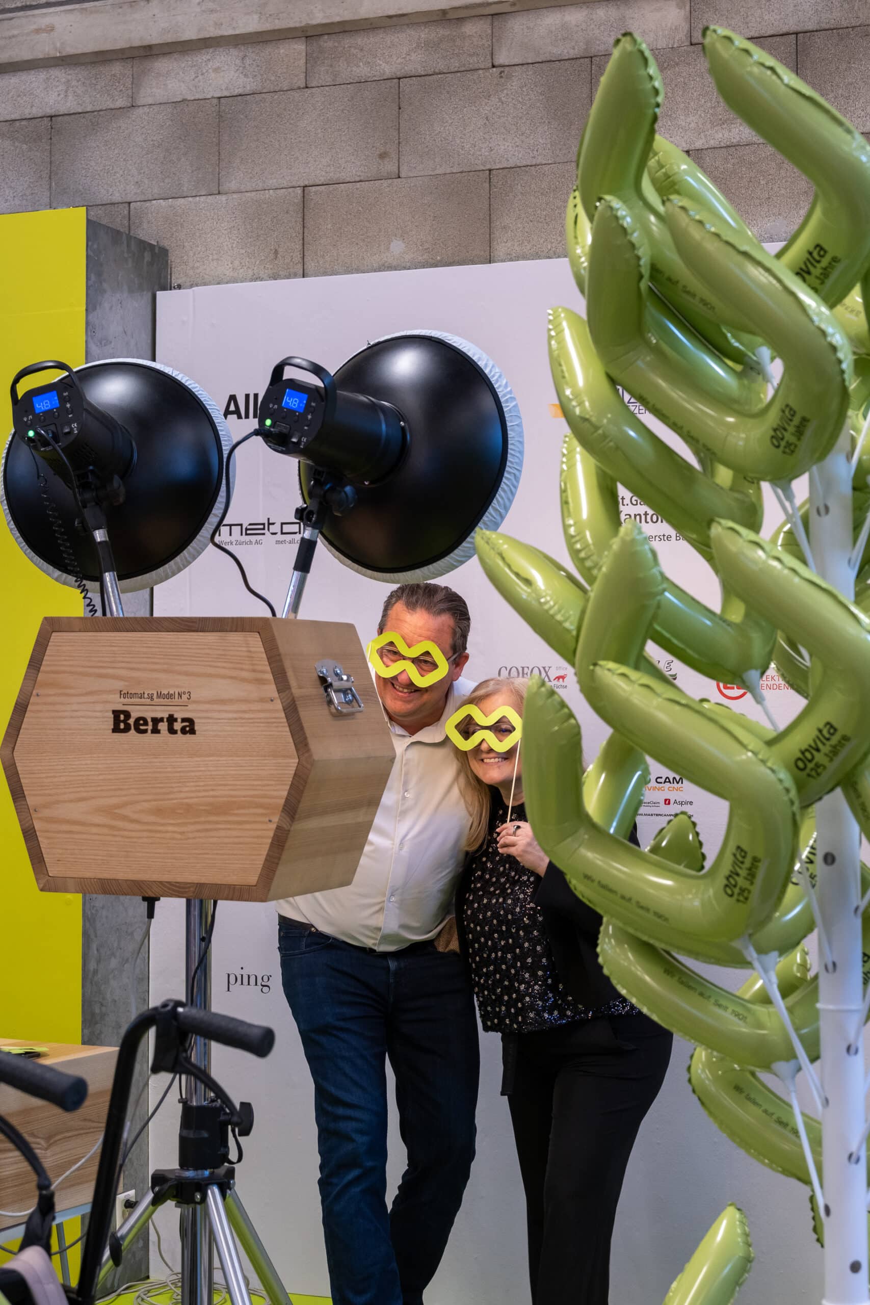 Ein Mann und eine Frau posieren zusammen an einem Fotoautomaten, beide tragen große gelbe Scherzbrillen. Im Vordergrund sind grüne Ballondekorationen zu sehen, und hinter ihnen ist eine professionelle Beleuchtungsanlage zu erkennen.