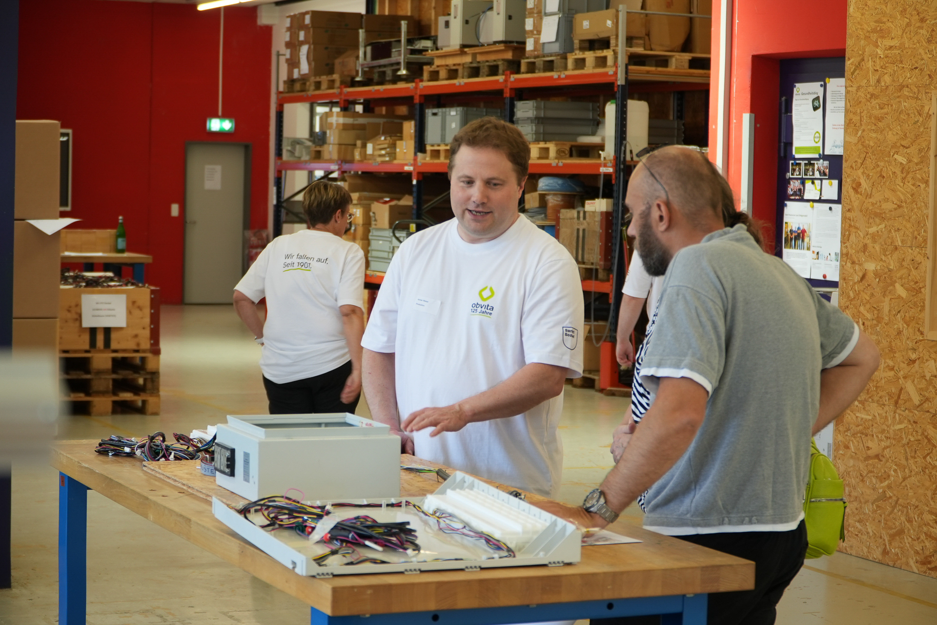 Zwei Männer stehen an einer Werkbank und unterhalten sich über Elektronikbauteile in einem Lager oder einer Werkstatt. Im Hintergrund gehen zwei Personen in weißen Hemden davon, und an den Wänden reihen sich Regale mit Kisten und Geräten aneinander.