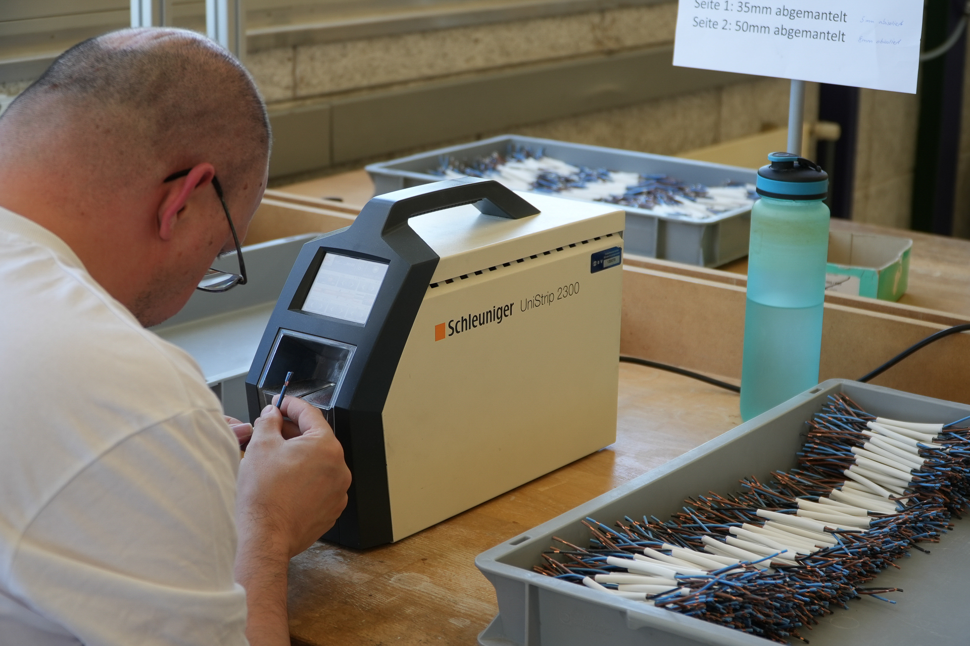 Eine Person bedient an einem Arbeitsplatz eine Schleuniger UniStrip 2300-Abisoliermaschine; daneben stehen Schalen mit vorbereiteten Kabeln und eine Wasserflasche. Über dem Arbeitsbereich sind Schilder mit Anweisungen angebracht.