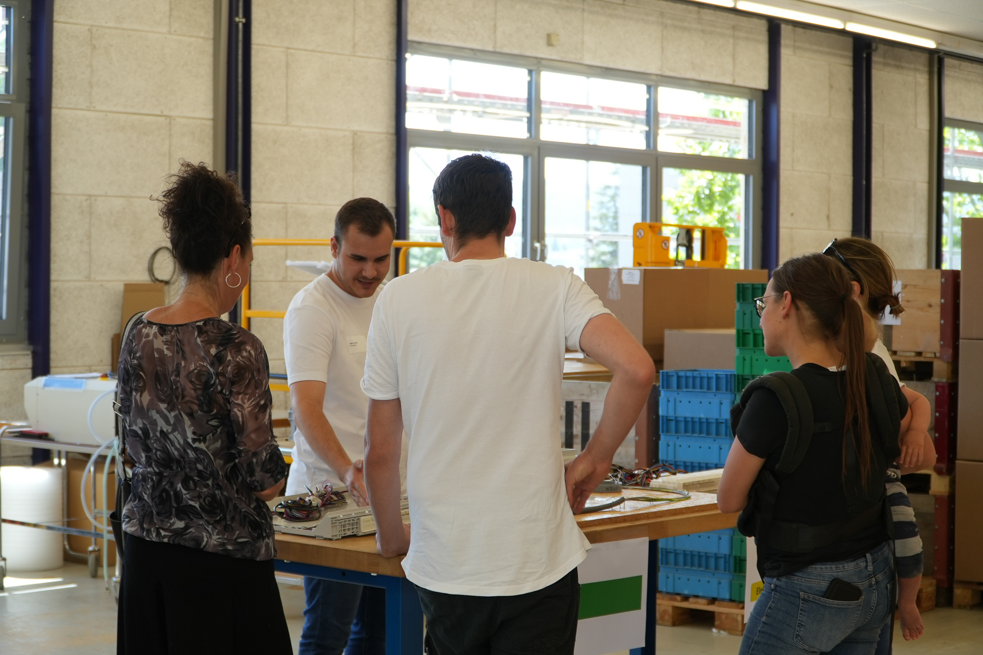 Vier Personen stehen in einem gut beleuchteten Raum um einen Tisch mit elektronischen Bauteilen herum und hören einem Mann zu, der etwas erklärt. Im Hintergrund sind Kisten und Geräte zu sehen, was auf eine Werkstatt oder ein Labor hindeutet.