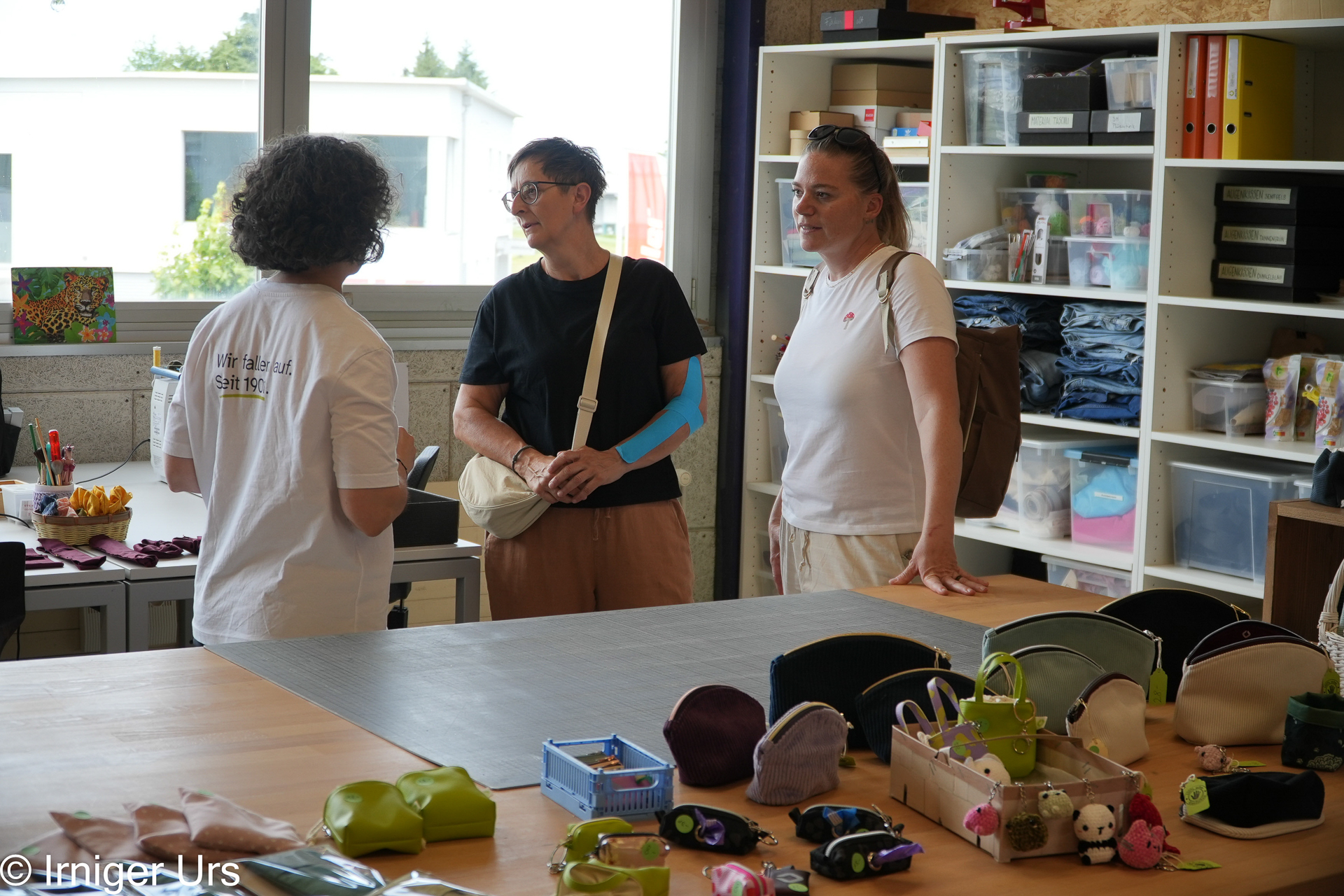 Drei Frauen stehen in einem hell beleuchteten Bastelraum und unterhalten sich. Im Vordergrund stehen Regale, Bastelmaterialien sowie verschiedene handgefertigte Beutel und Spielzeuge, die auf einem großen Tisch ausgestellt sind.