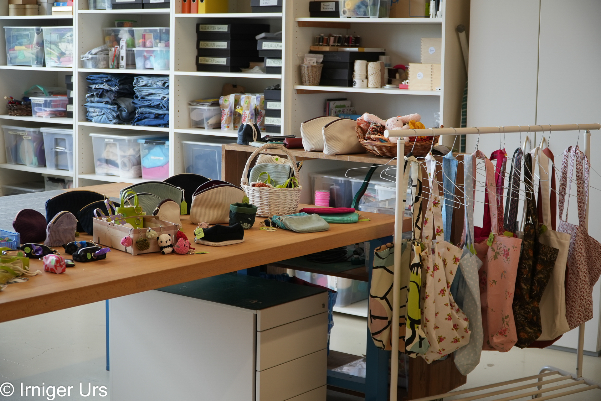 Ein Bastelatelier mit Regalen voller Stoffe und Bastelmaterialien, einem Tisch, auf dem handgefertigte Beutel und kleines Spielzeug ausgestellt sind, sowie einem Ständer mit bunten Taschen und Geldbörsen.
