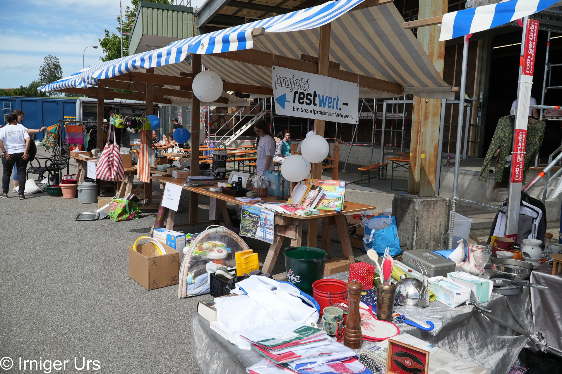 An einem Flohmarktstand auf dem Bürgersteig werden unter einem gestreiften Vordach verschiedene Secondhand-Artikel wie Spielzeug, Küchenutensilien und Bücher angeboten. Hinter dem Tisch stehen zwei Personen, über ihnen hängt ein Schild mit der Aufschrift „Projekt Restwert“.