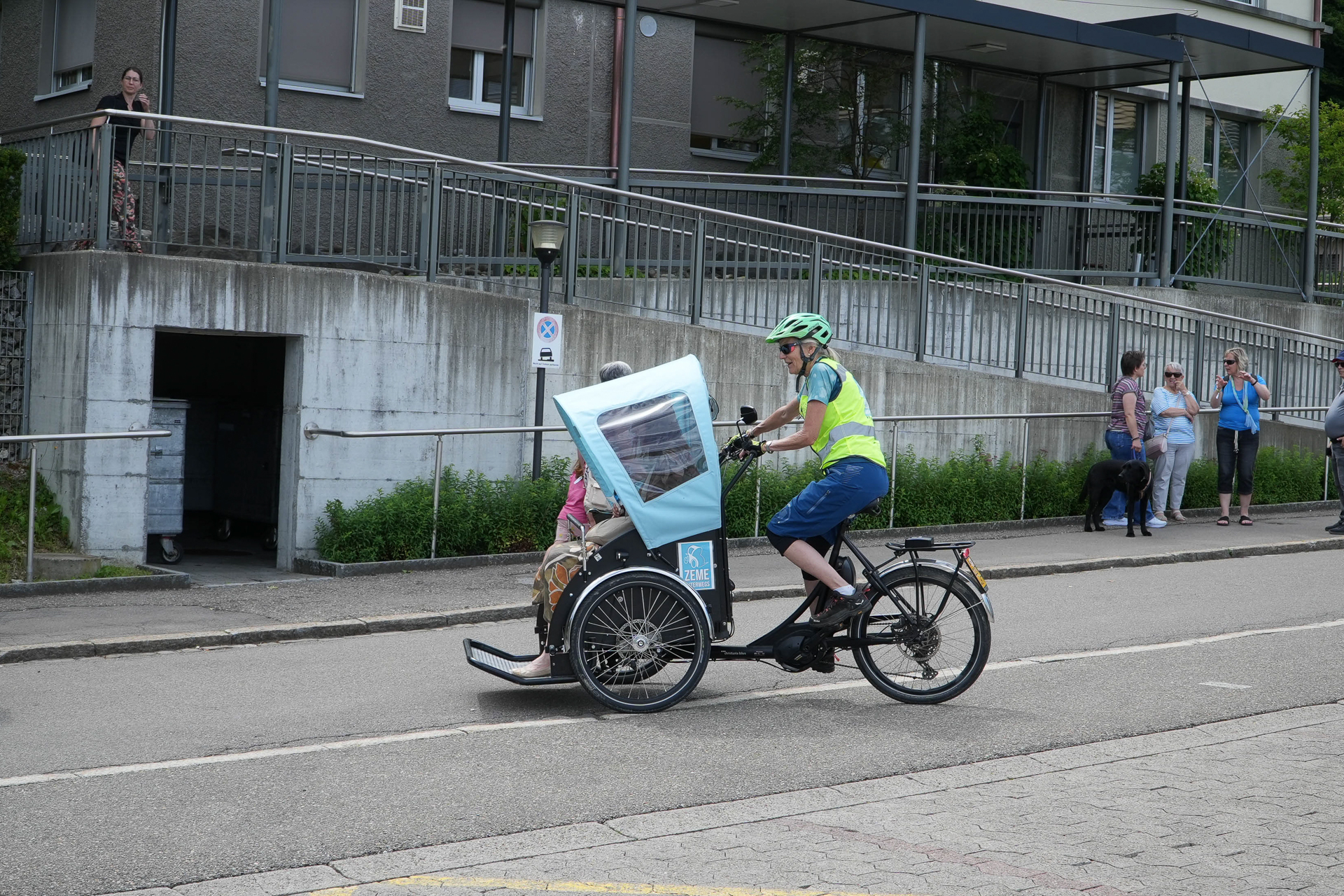 Eine Person, die einen Helm und eine Warnweste trägt, fährt mit einem Dreirad mit überdachtem Beifahrersitz, auf dem eine ältere Person sitzt, auf einer Straße in der Nähe eines Gebäudes, vor dem Menschen stehen.