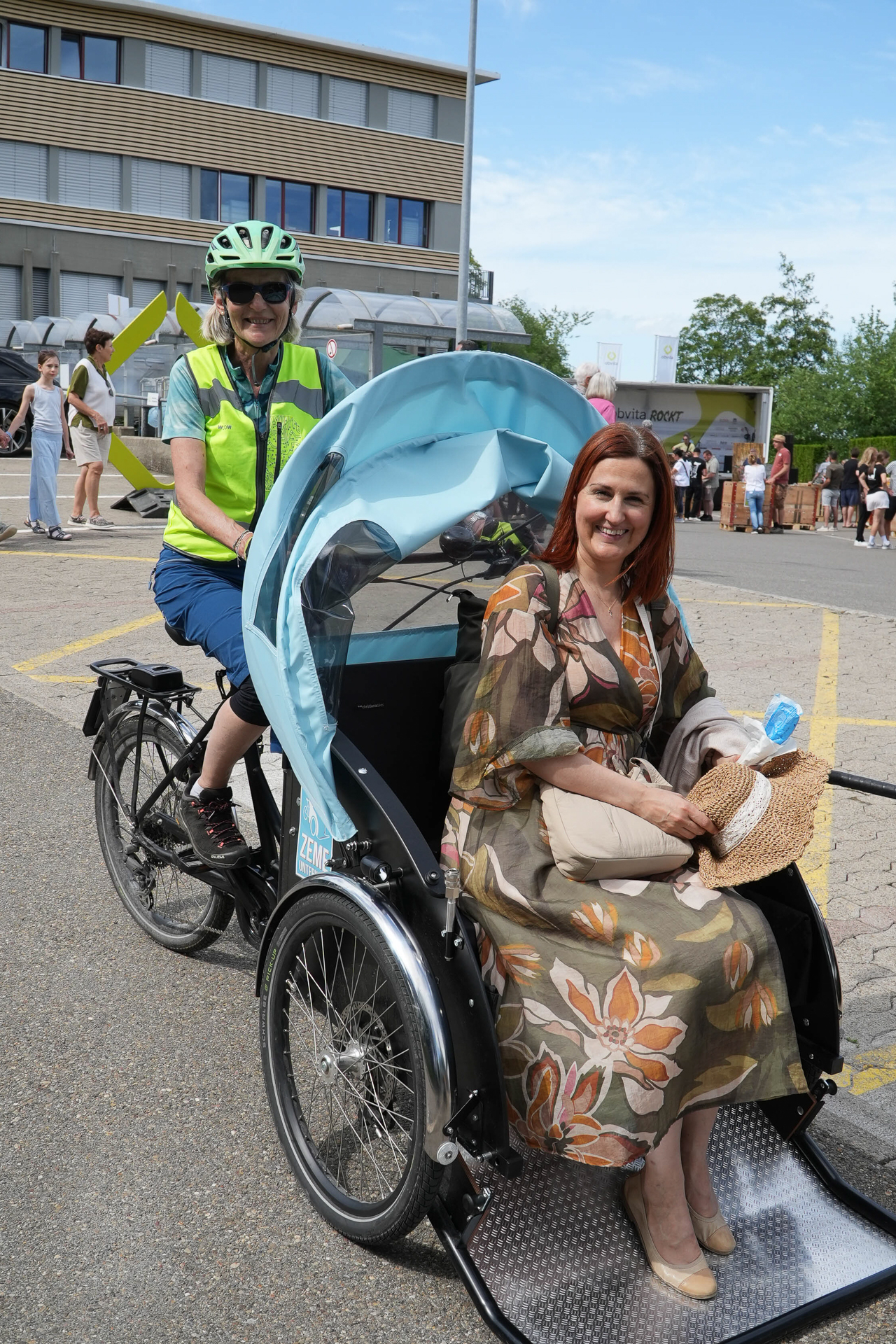 Eine Frau in einem geblümten Kleid sitzt lächelnd auf dem Beifahrersitz eines Fahrrads im Rikscha-Stil, während eine andere Frau mit Sicherheitsweste und Helm das Fahrrad fährt. Im Hintergrund sind an einem sonnigen Tag Menschen und Gebäude zu sehen.