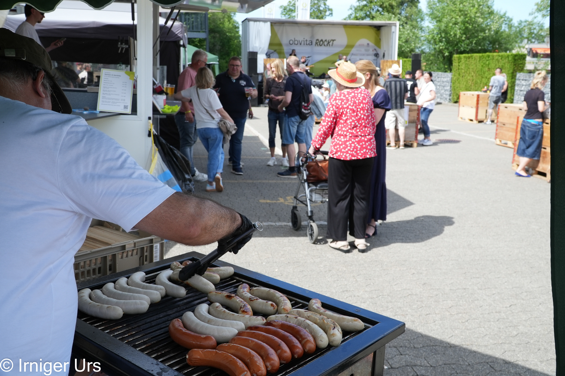 An einem sonnigen Tag grillt jemand auf einem Markt im Freien verschiedene Würstchen, während sich im Hintergrund Menschen versammeln und unterhalten.