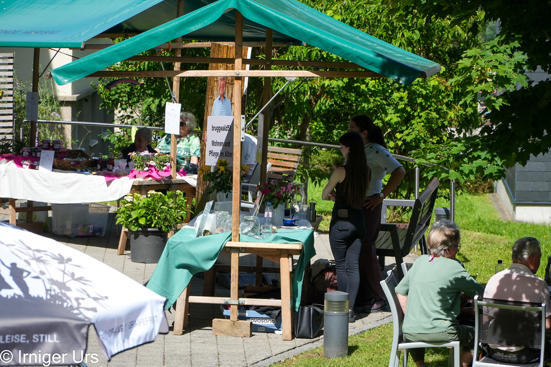 Ein kleiner Markt im Freien, auf dem Menschen in der Nähe von Holzständen unter grünen Markisen sitzen und stehen, während auf den Tischen Pflanzen und Waren ausgestellt sind und das Sonnenlicht durch die Bäume fällt.