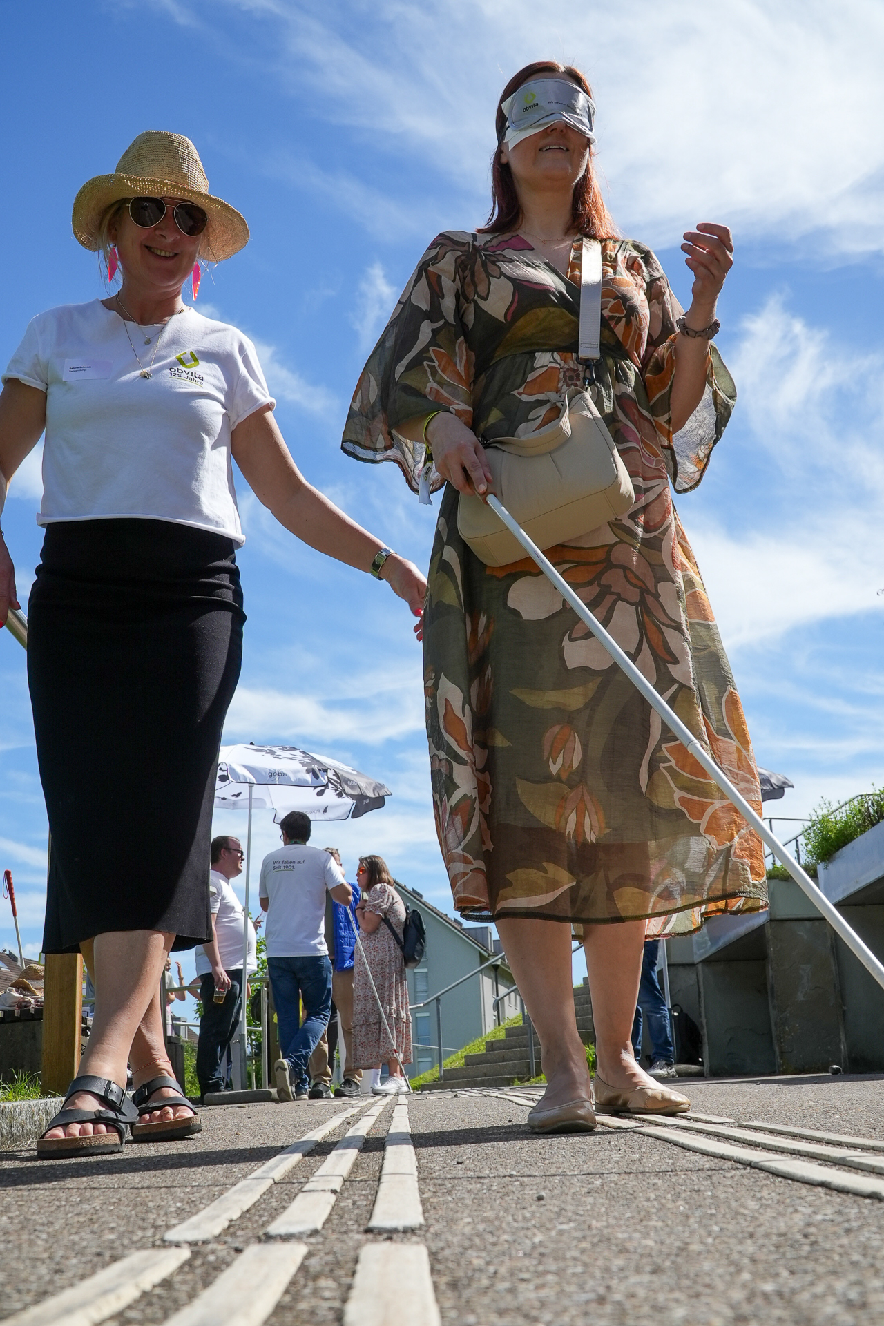 Eine Frau mit einer Augenbinde und einem weißen Blindenstock geht auf einem taktilen Pflaster entlang, geführt von einer anderen lächelnden Frau mit Hut bei einer Veranstaltung im Freien unter blauem Himmel. Im Hintergrund sind Menschen und Regenschirme zu sehen.