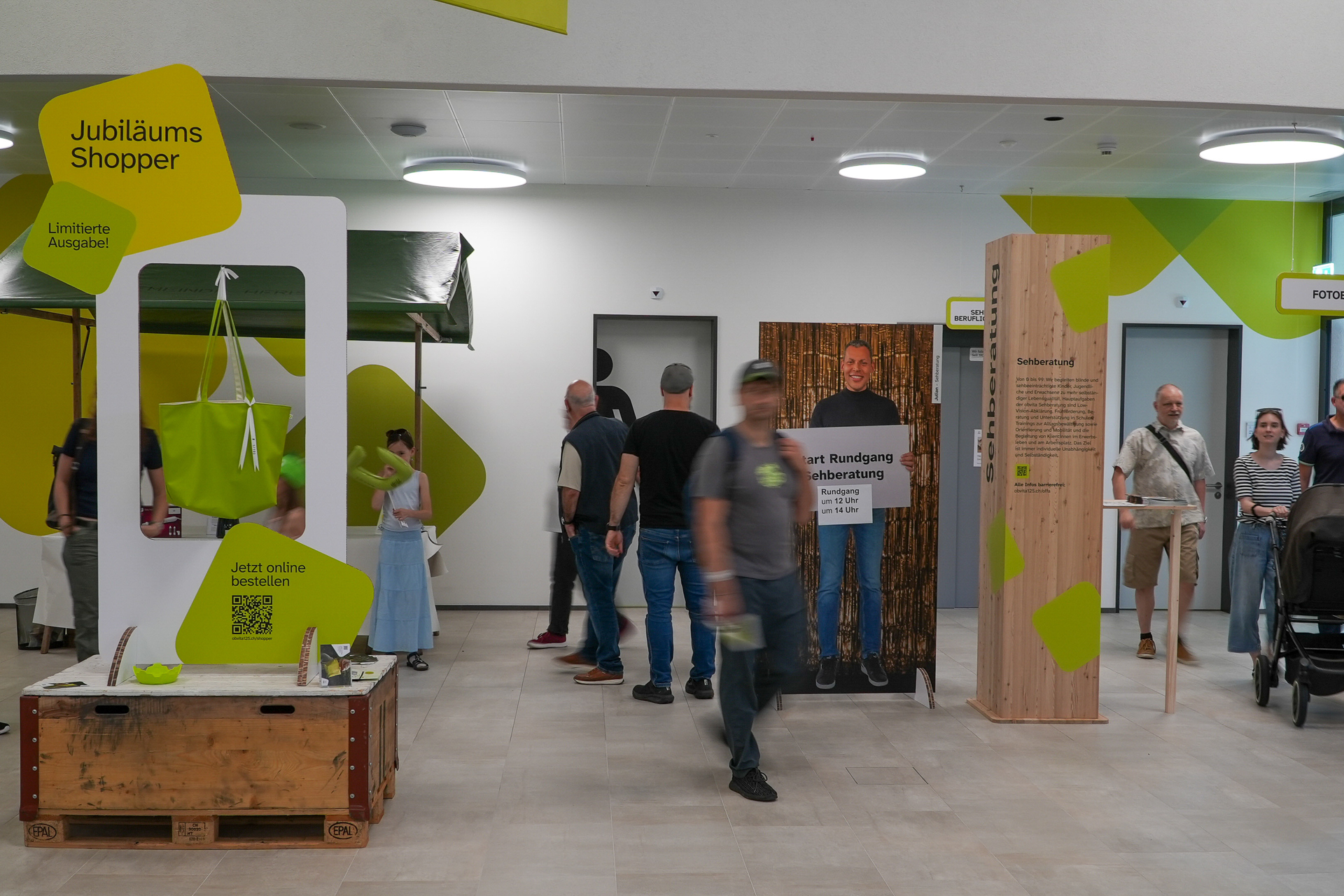 Menschen gehen durch ein modernes Gebäude mit Ausstellungsstücken, darunter eine grüne Tasche auf einem Ständer mit der Aufschrift „Jubiläums Shopper“ und eine Holzfigur, die als Fotomotiv dient. Einige stehen still, andere sind in Bewegung.