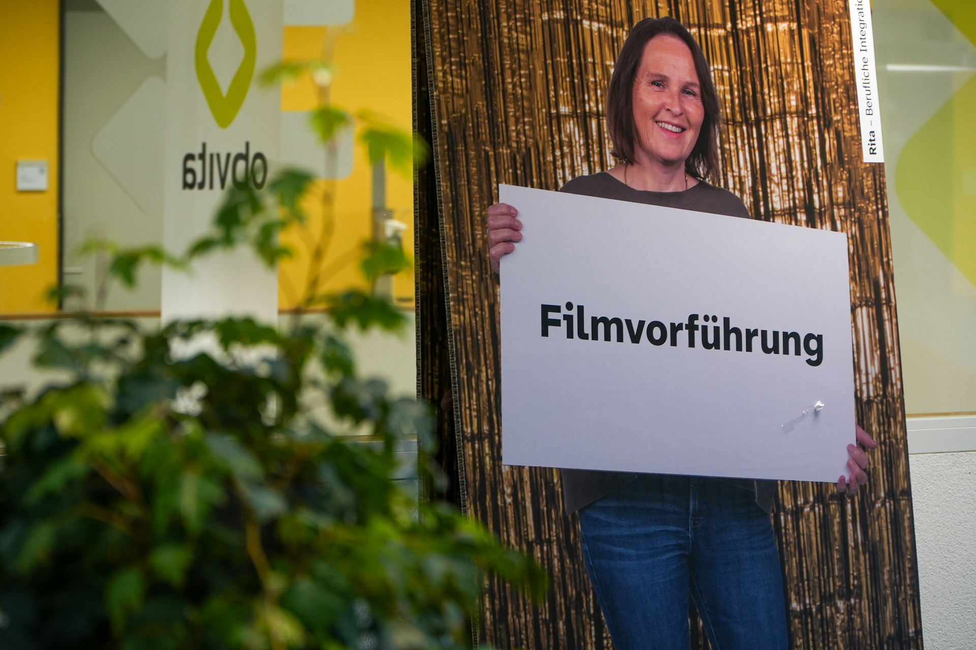 Eine Frau hält lächelnd ein Schild mit der Aufschrift „Filmvorführung“ in der Hand. Sie steht vor einem Hintergrund mit Bambusmuster. Links sind grünes Laub und eine Glaswand mit der Aufschrift „obvia“ zu sehen.