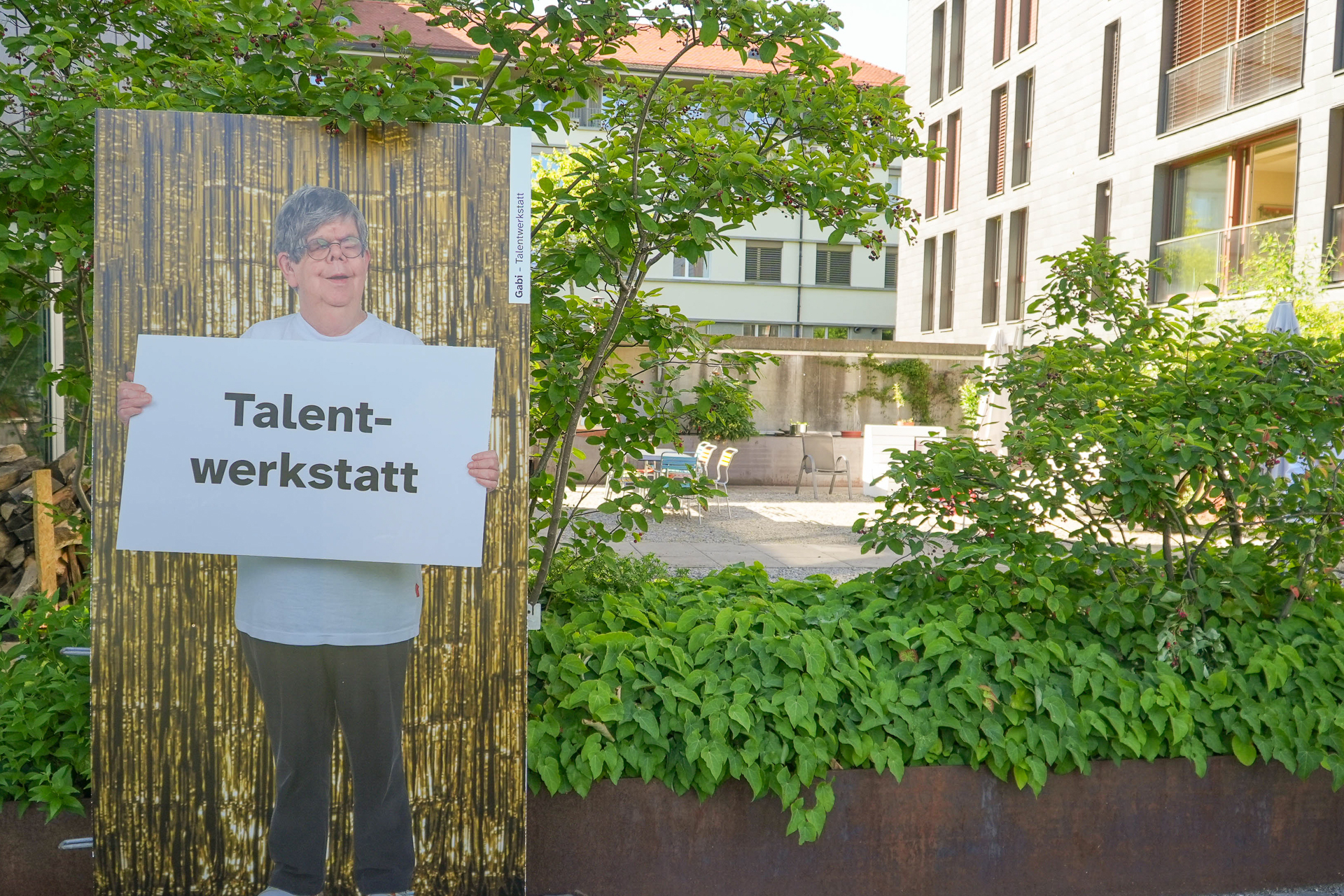 Eine Person steht im Freien vor einem goldenen Vorhang als Kulisse und hält ein Schild mit der Aufschrift „Talent-werkstatt“ in der Hand; im Hintergrund sind Grünflächen und moderne Gebäude zu sehen.