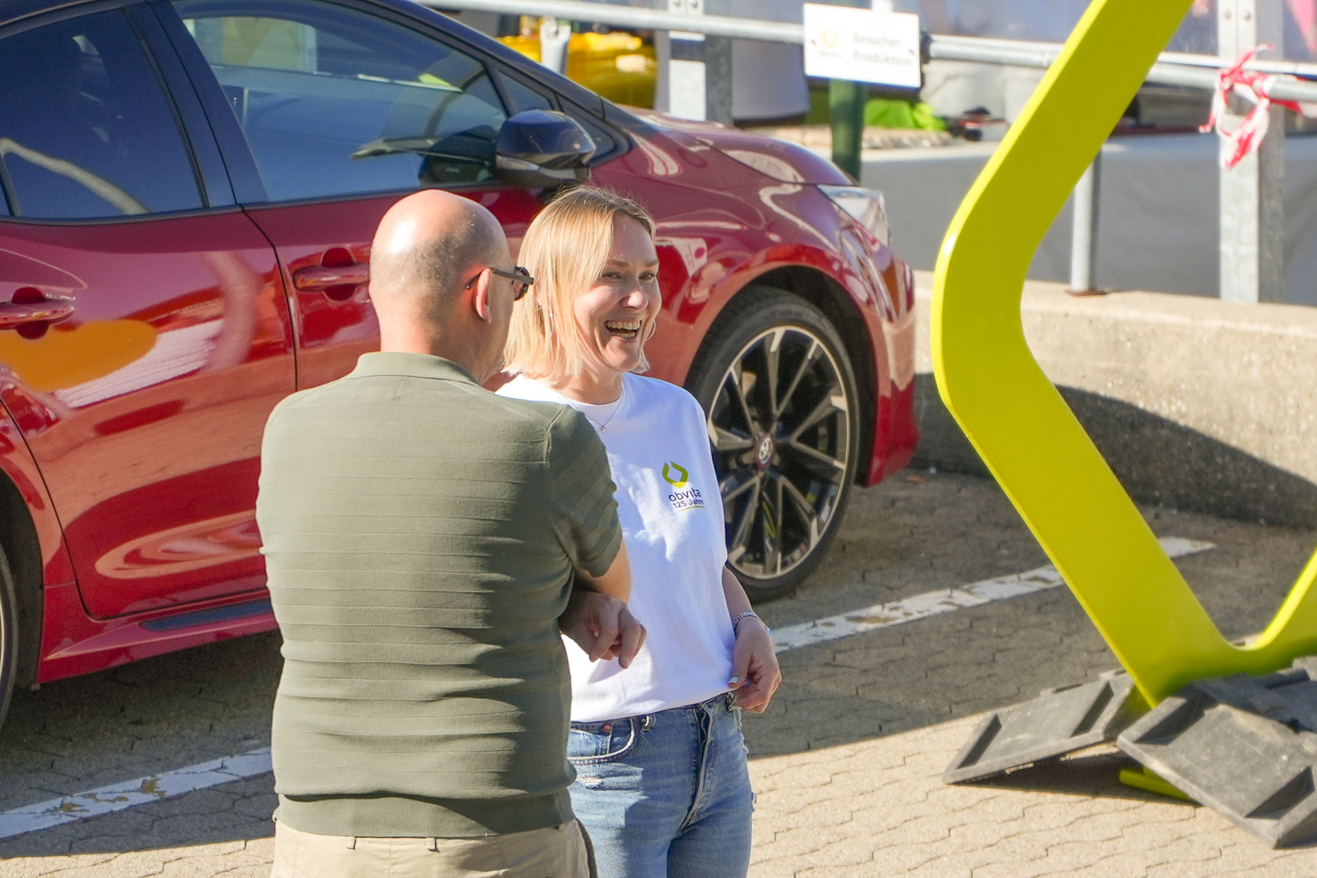 Ein Mann und eine Frau stehen lächelnd und unterhalten sich auf einem sonnigen Parkplatz neben einem roten Auto und einem gelben Gebäude. Die Frau trägt ein weißes Shirt mit einem bunten Logo; der Mann trägt ein grünes Polohemd.