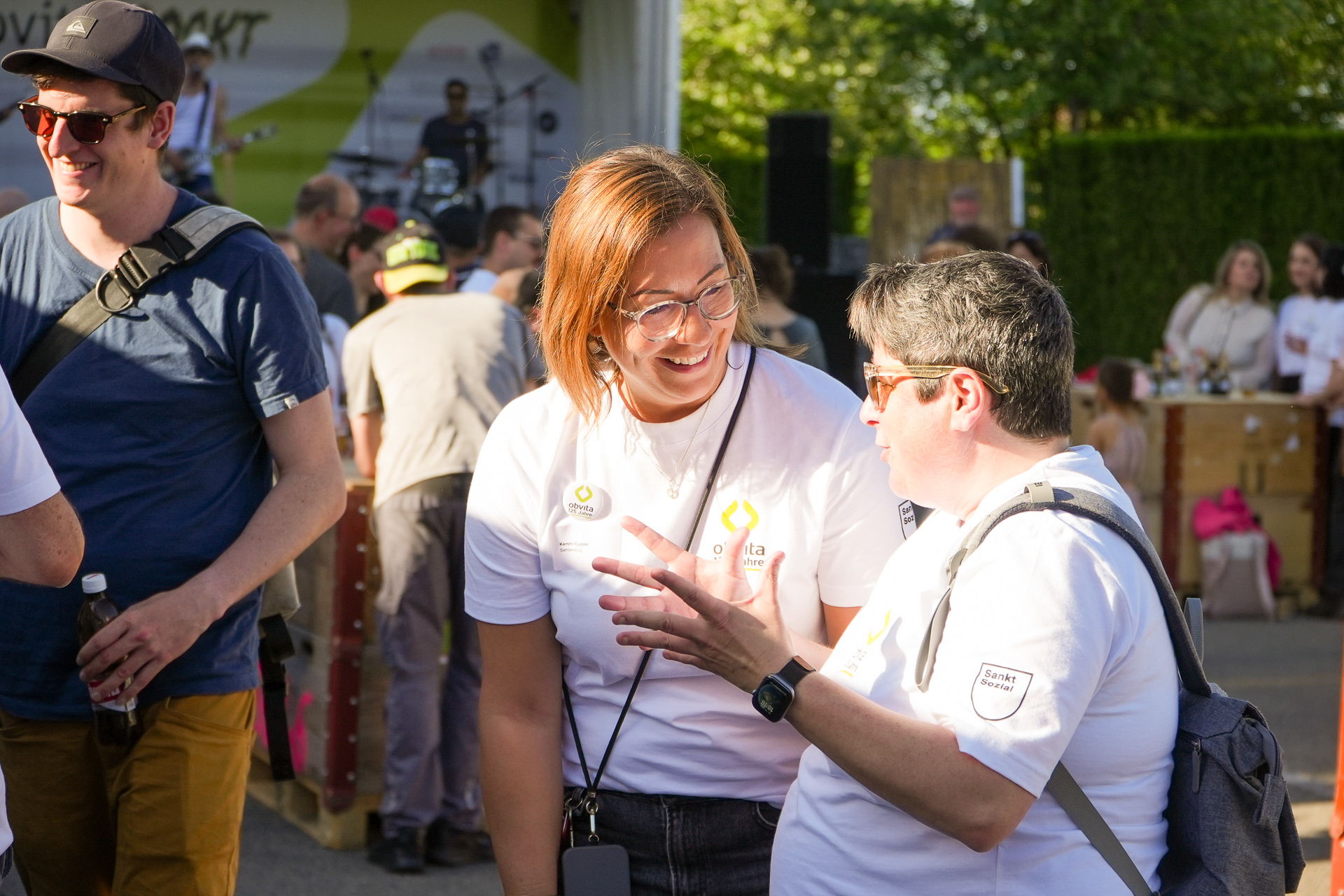 Zwei Personen in weißen Hemden und mit Namensschildern lächeln und unterhalten sich bei einer Veranstaltung im Freien mit anderen Menschen; im Hintergrund ist eine Bühne zu sehen. Sonnenlicht fällt auf sie, und in der Ferne sind Bäume zu erkennen.