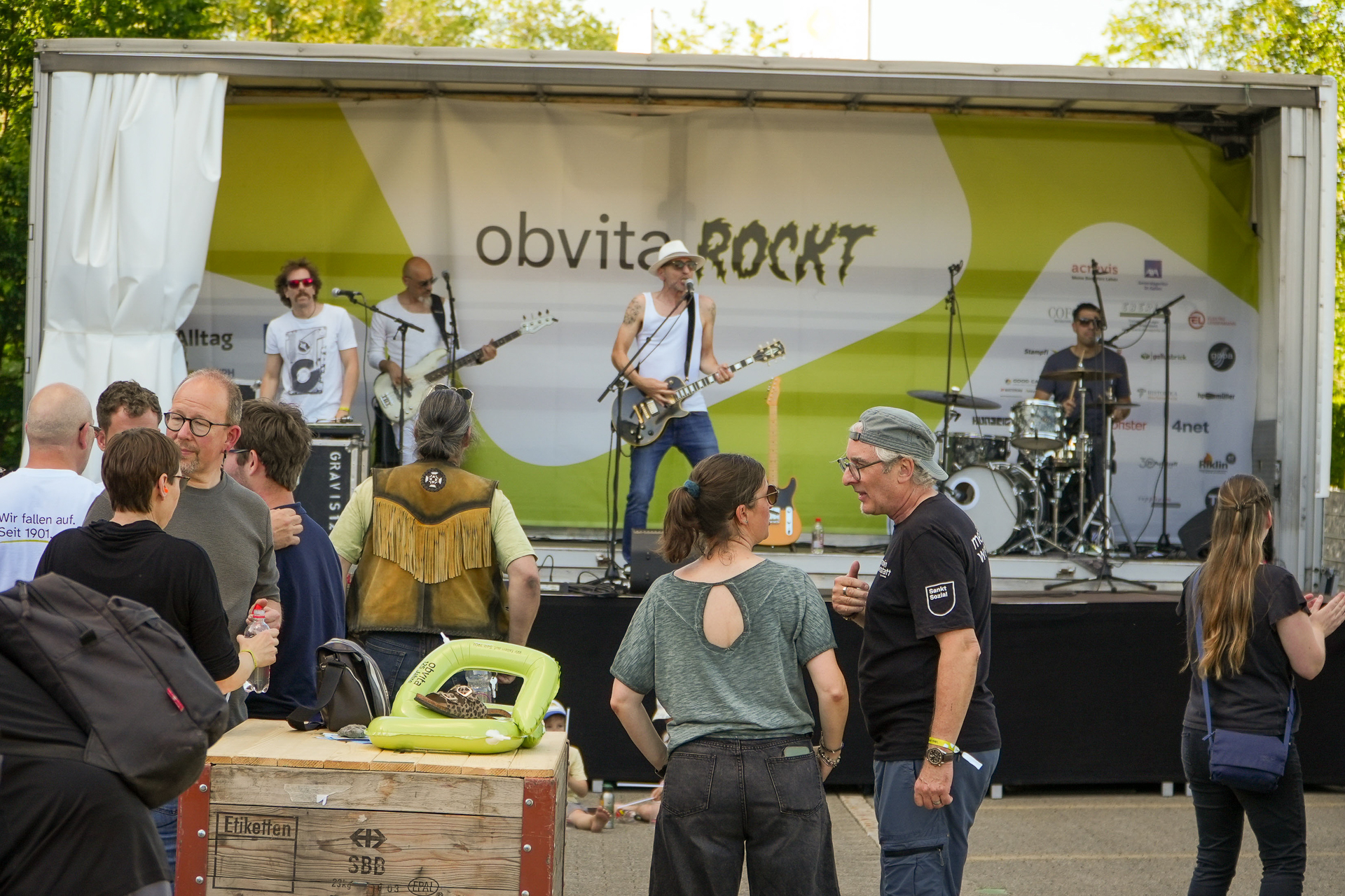 Eine Rockband tritt auf einer mit „obvita ROCKT“ beschrifteten Außenbühne auf, während eine Menschenmenge an einem sonnigen Tag zuschaut, sich unterhält und die Veranstaltung genießt. Musikinstrumente und Logos der Veranstaltungssponsoren sind zu sehen.
