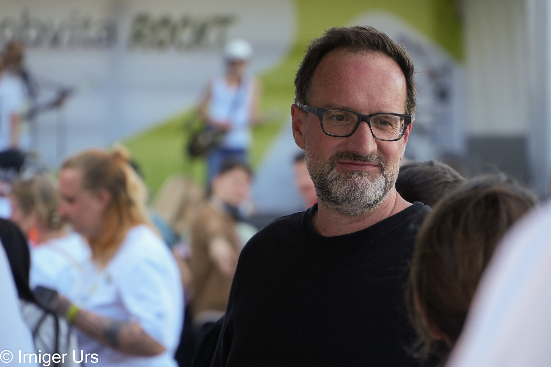 Ein bärtiger Mann mit Brille steht inmitten einer Menschenmenge bei einer Veranstaltung im Freien oder einem Festival, lächelt und blickt zur Seite. Im Hintergrund sind Menschen und eine Bühne mit Künstlern zu sehen.
