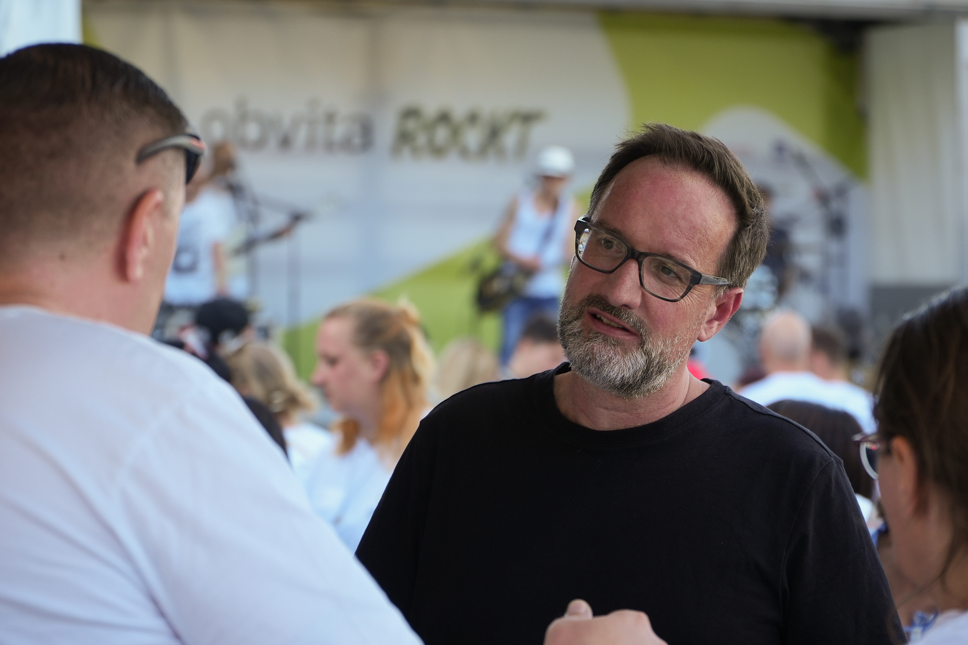 Ein Mann mit Brille und schwarzem Hemd unterhält sich bei einer Veranstaltung im Freien mit einem anderen Mann, im Hintergrund sind Menschen und eine Bühne zu sehen. Auf dem Banner auf der Bühne ist das Wort „ROCKT“ teilweise zu erkennen.