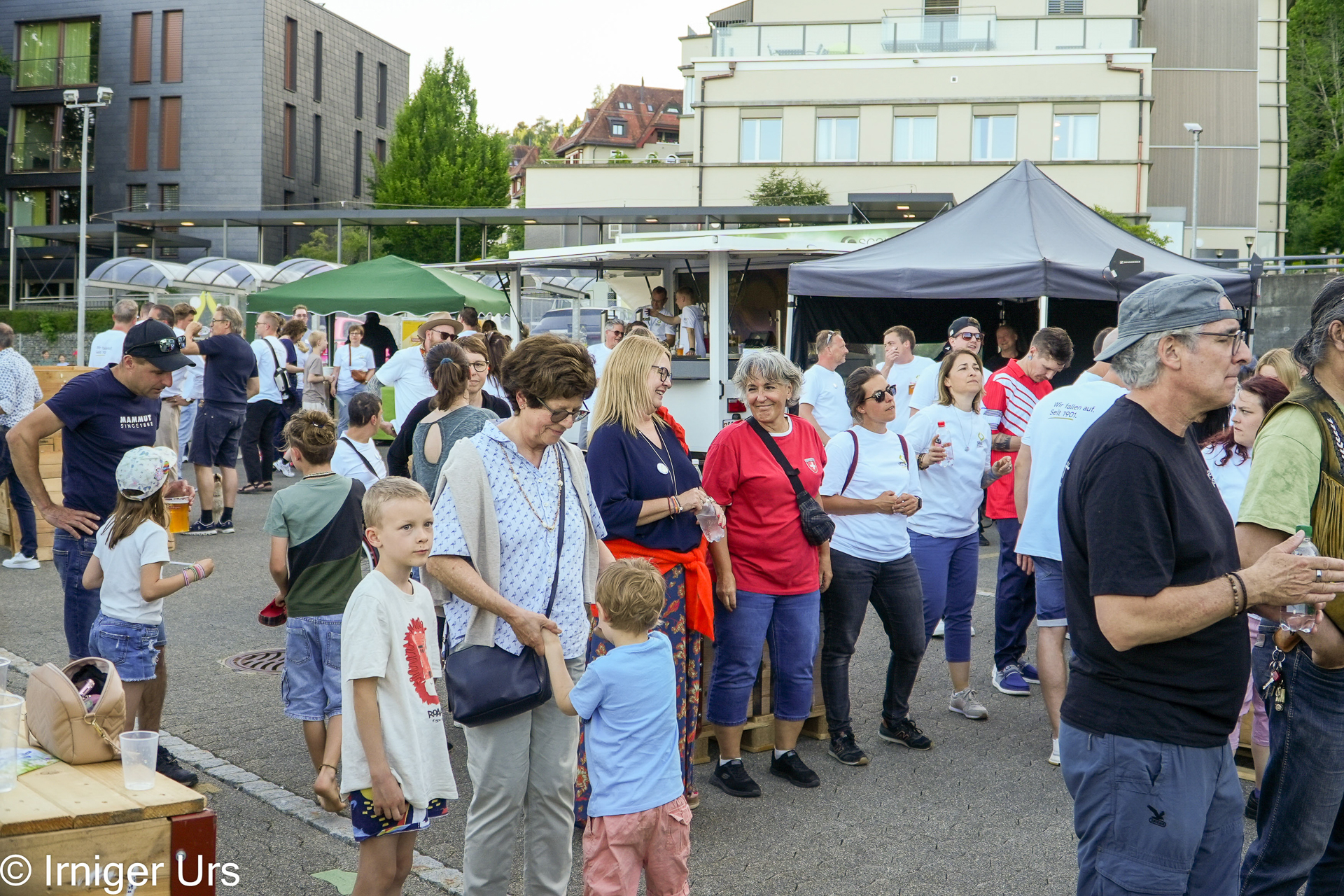 An einem sonnigen Tag versammelt sich eine Gruppe von Menschen, darunter Erwachsene und Kinder, im Freien bei einer Veranstaltung, bei der im Hintergrund Imbissstände und Zelte zu sehen sind. Einige tragen rote oder blaue Hemden und scheinen sich zu unterhalten.
