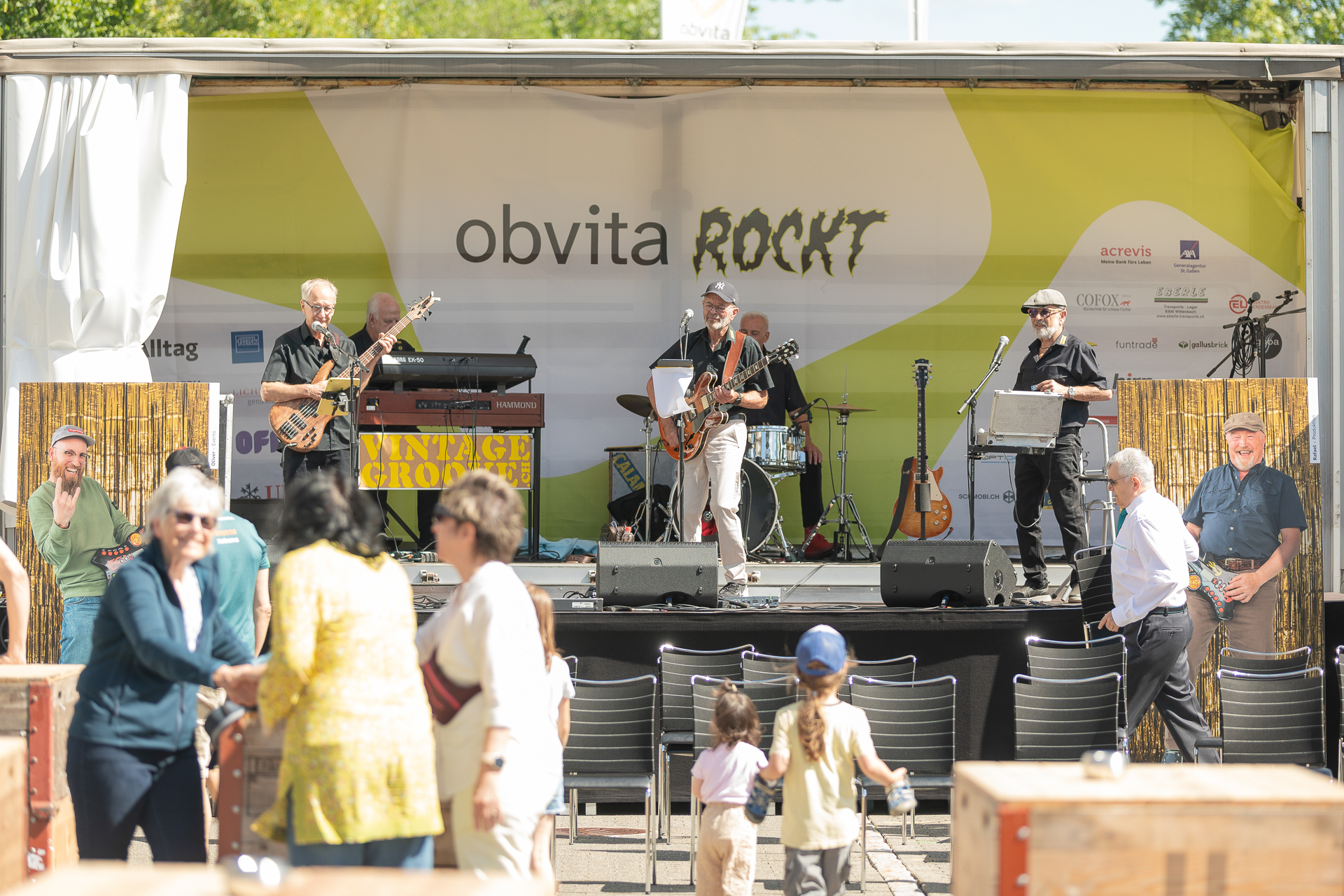 Eine Band namens „Vintage Groove“ tritt auf einer Open-Air-Bühne im Rahmen der Veranstaltung „obvita ROCKT“ auf, während Menschen jeden Alters an diesem sonnigen Tag vor der Bühne zuschauen, sich unterhalten und tanzen.
