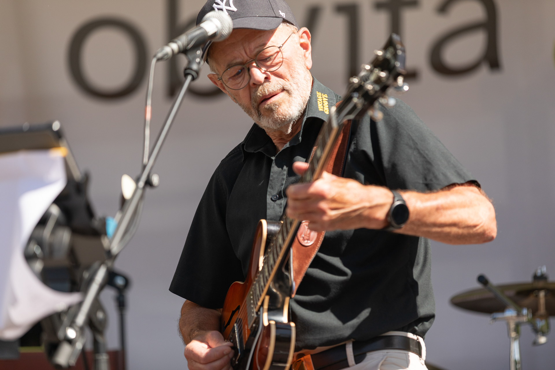 Ein älterer Mann mit Brille, schwarzem Hemd und Baseballmütze spielt auf einer Außenbühne E-Gitarre und singt ins Mikrofon, im Hintergrund sind verschwommene Musikinstrumente und ein Schlagzeug zu sehen.