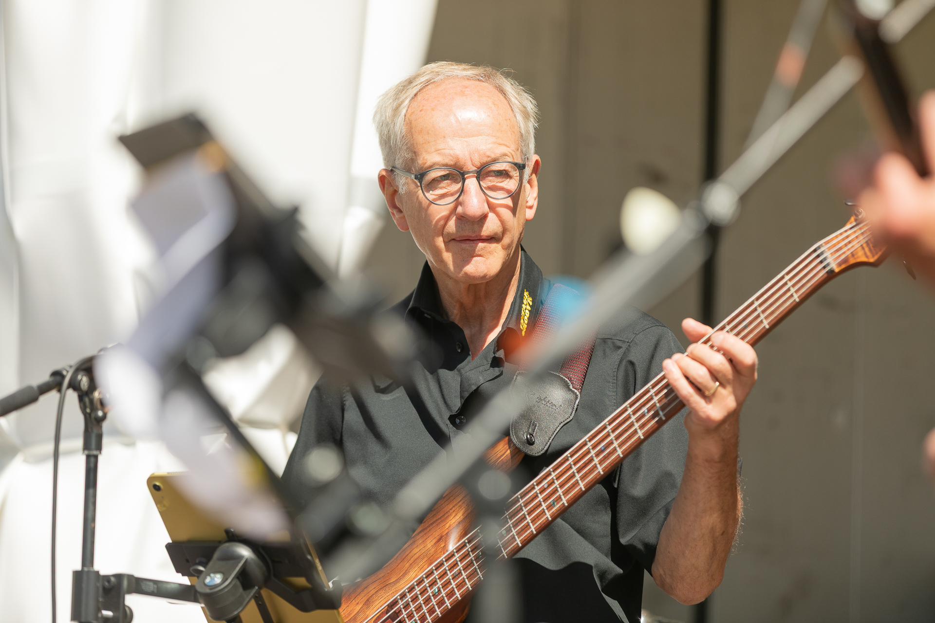 Ein älterer Mann mit Brille und schwarzem Hemd spielt auf der Bühne E-Bass, während Sonnenlicht auf ihn fällt und im Vordergrund die Ausrüstung zu sehen ist.