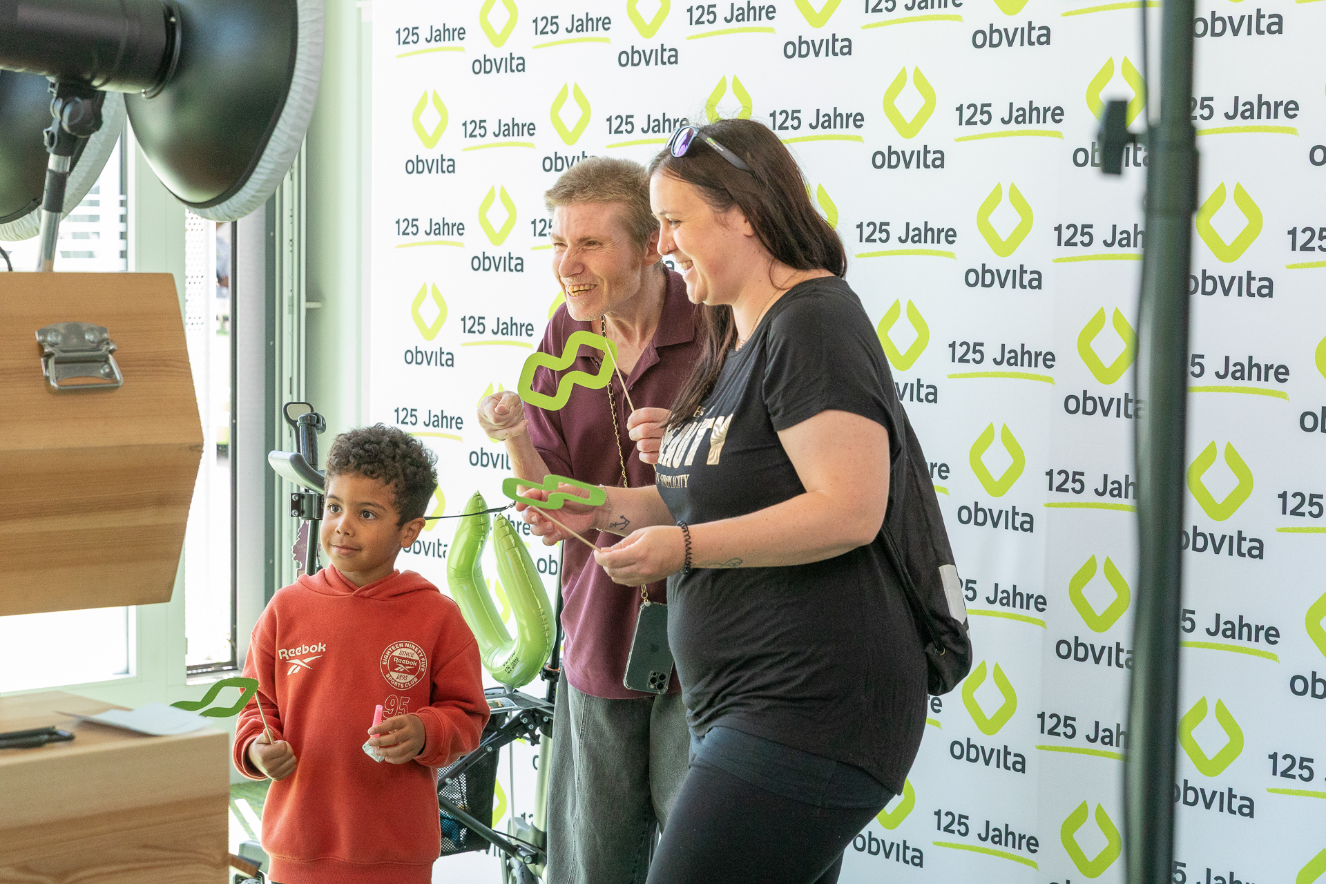 Drei Personen, darunter ein Kind in einem roten Sweatshirt, posieren und lächeln für ein Foto vor einem Hintergrund mit den Aufschriften „obvita“ und „125 Jahre“. Sie halten grüne Requisiten in Form des obvita-Logos in den Händen.