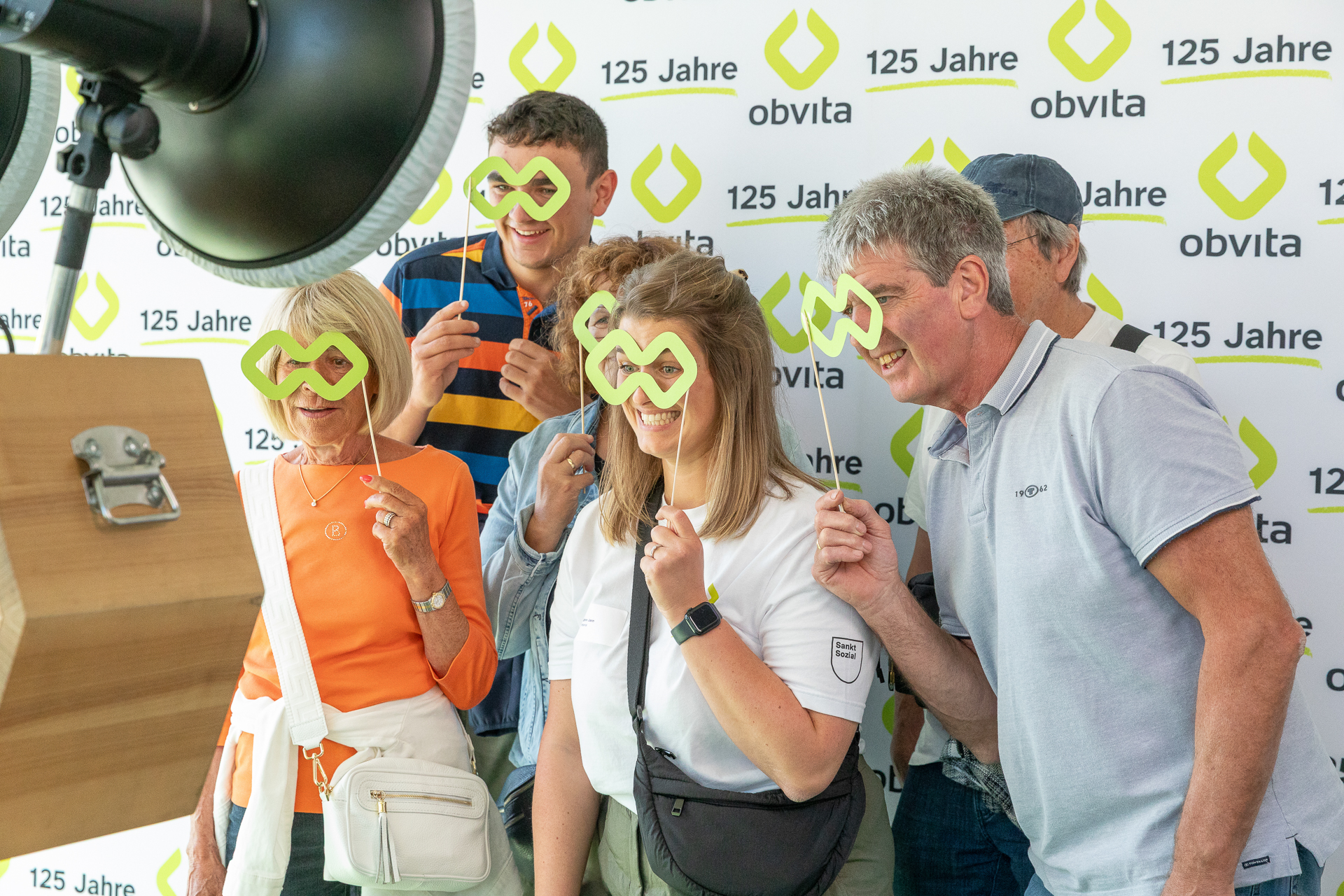 Eine Gruppe lächelnder Menschen, die Spielzeugbrillen tragen, posiert gemeinsam in einem Raum für ein lustiges Foto. Ein Mann in einem blauen Hemd umarmt eine Frau in Weiß, während sich andere um sie herum versammeln. Im Hintergrund sind Werbebanner und ein Fenster zu sehen.