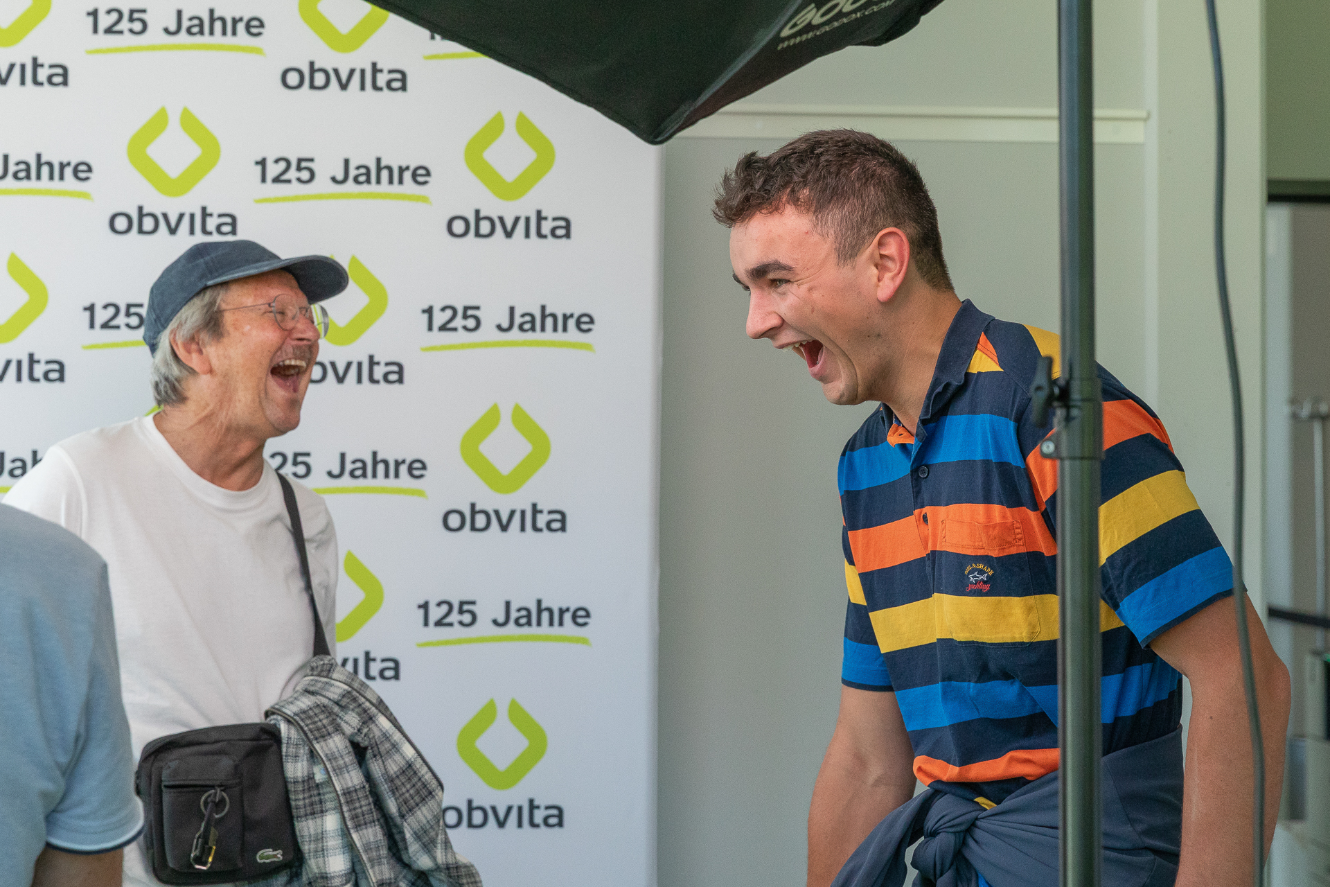 Zwei Männer lachen gemeinsam vor einem Hintergrund, auf dem „obvita 125 Jahre“ zu lesen ist. Der eine trägt ein gestreiftes Hemd, der andere ein weißes Hemd und eine Mütze. Die Stimmung ist fröhlich und lebhaft.