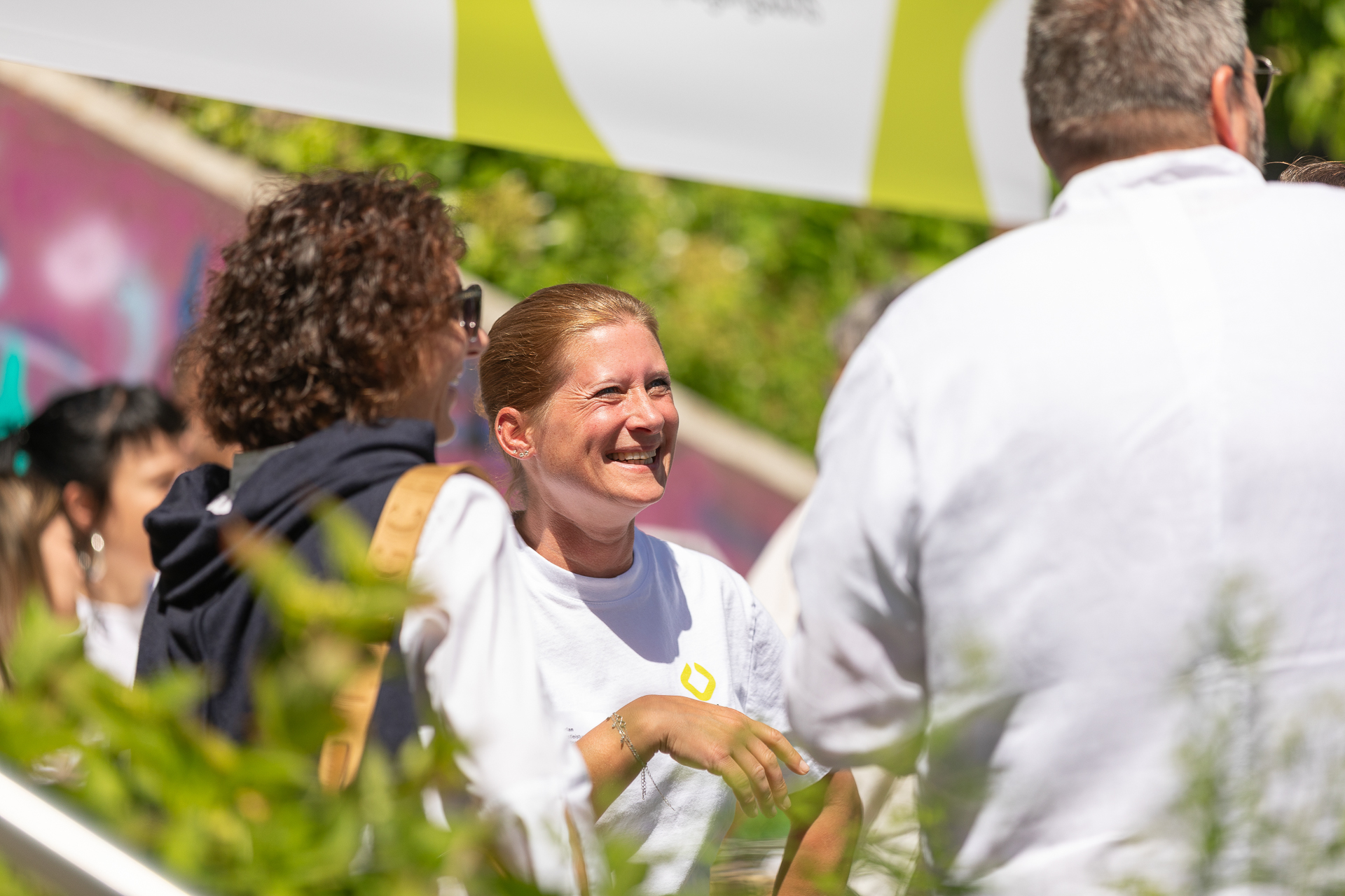 Eine Frau in einem weißen Hemd lächelt und unterhält sich an einem sonnigen Tag bei einer Veranstaltung im Freien mit anderen. Im Hintergrund sind Grünflächen und verschwommene Menschen zu sehen.