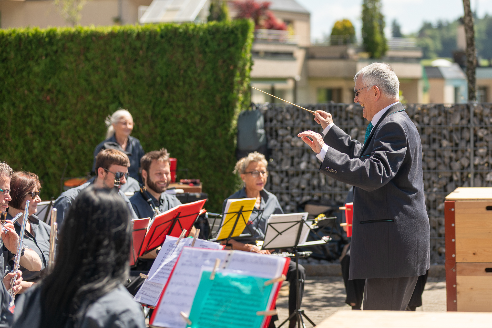 Ein Dirigent leitet ein Orchester im Freien, dessen Musiker sitzen und verschiedene Instrumente spielen. Man sieht Notenständer mit bunten Notenblättern, im Hintergrund sind Grünflächen und Gebäude zu sehen.