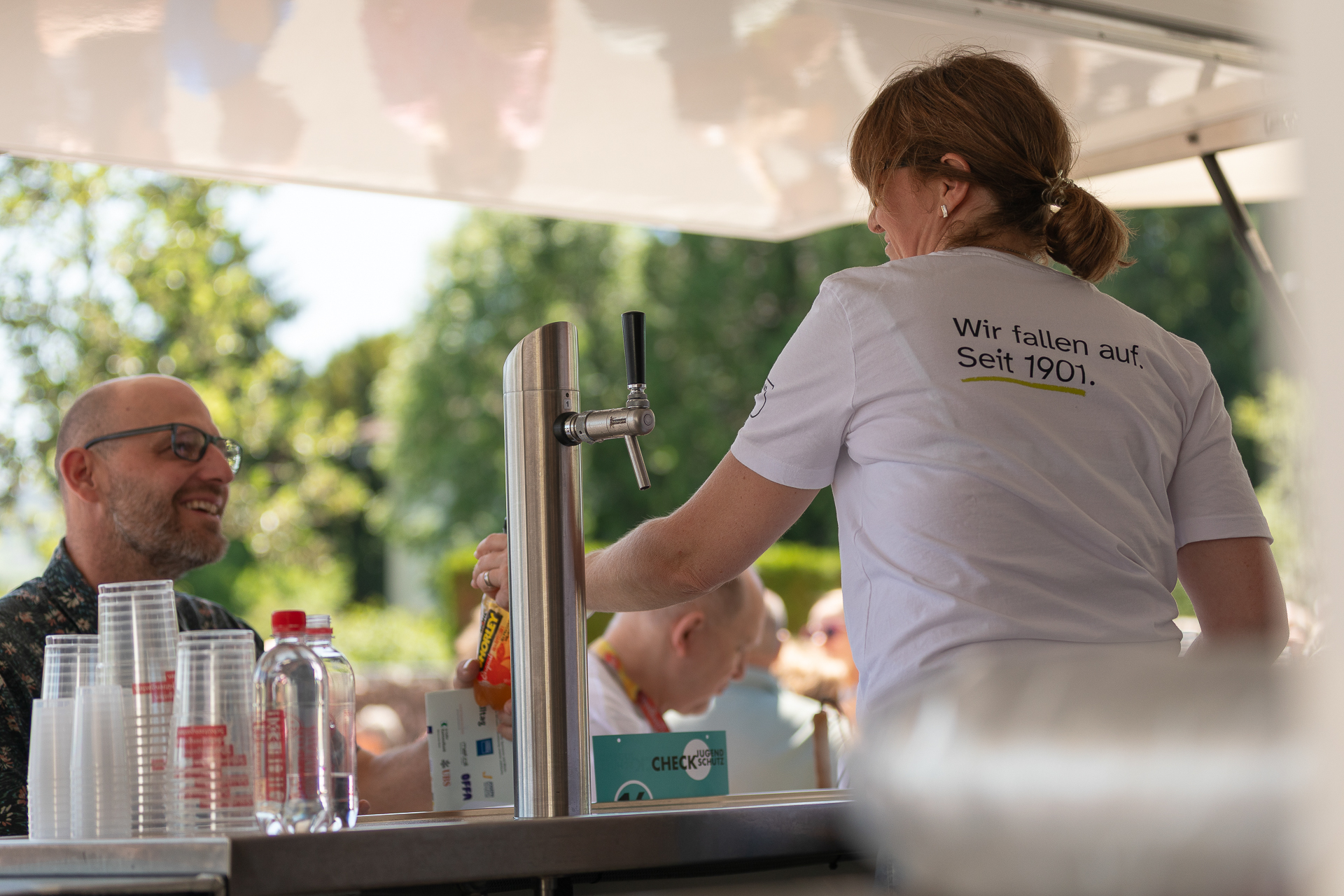 Eine Frau in einem weißen Hemd mit deutscher Aufschrift serviert bei einer Veranstaltung im Freien Getränke und lächelt einen Mann auf der anderen Seite der Theke an. Auf der Theke sind Stapel von Plastikbechern, Flaschen und ein Getränkehahn zu sehen. Im Hintergrund sind Bäume zu erkennen.