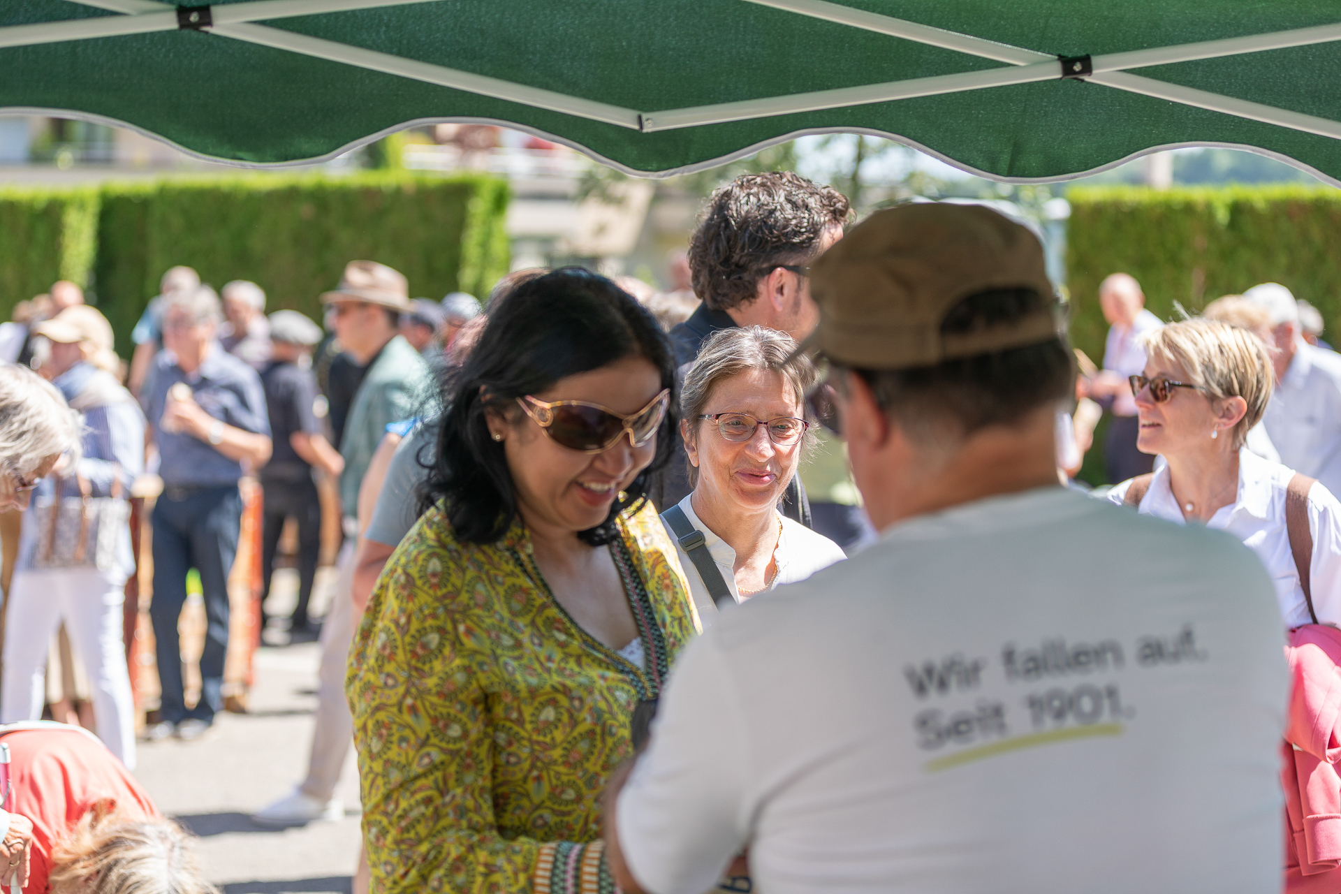 An einem sonnigen Tag versammeln sich Menschen im Freien unter einem grünen Blätterdach und unterhalten sich. Im Vordergrund steht eine lächelnde Frau in gemusterter Kleidung, im Hintergrund sind weitere Menschen zu sehen, die sich unterhalten.