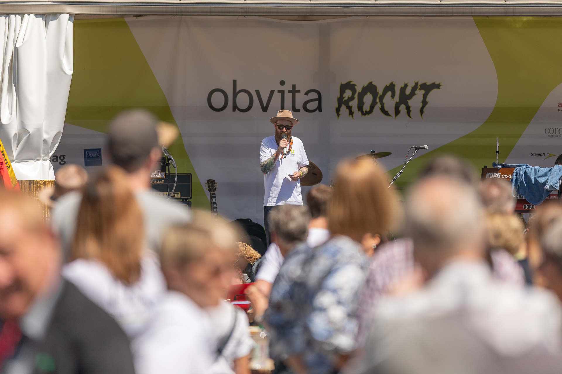 Ein Mann mit Hut und Sonnenbrille spricht auf einer Außenbühne, über der ein gelb-weißes Banner mit der Aufschrift „obvita ROCKT“ hängt, in ein Mikrofon, während im Vordergrund eine Menschenmenge zusieht.