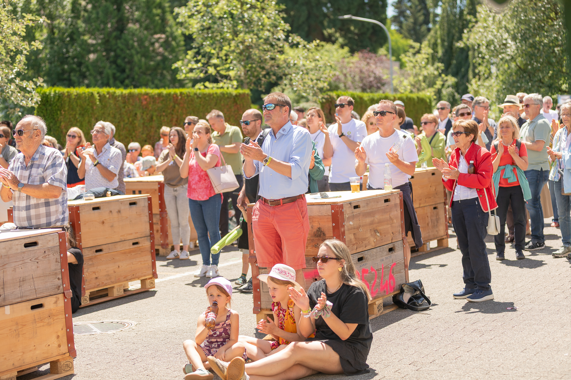 An einem sonnigen Tag steht und sitzt eine bunte Gruppe von Menschen im Freien, klatscht und verfolgt eine Veranstaltung. Einige sitzen auf dem Bürgersteig, während andere hinter Holzkisten stehen; im Hintergrund sind Bäume und Grünflächen zu sehen.