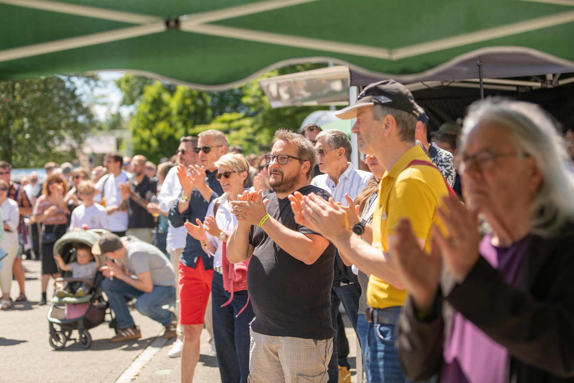 Eine bunte Gruppe von Menschen steht an einem sonnigen Tag im Freien unter einem Zelt, klatscht und verfolgt eine Veranstaltung. Einige tragen Sonnenbrillen und Freizeitkleidung; in der Menge sind Erwachsene, Kinder und ein Kinderwagen zu sehen.