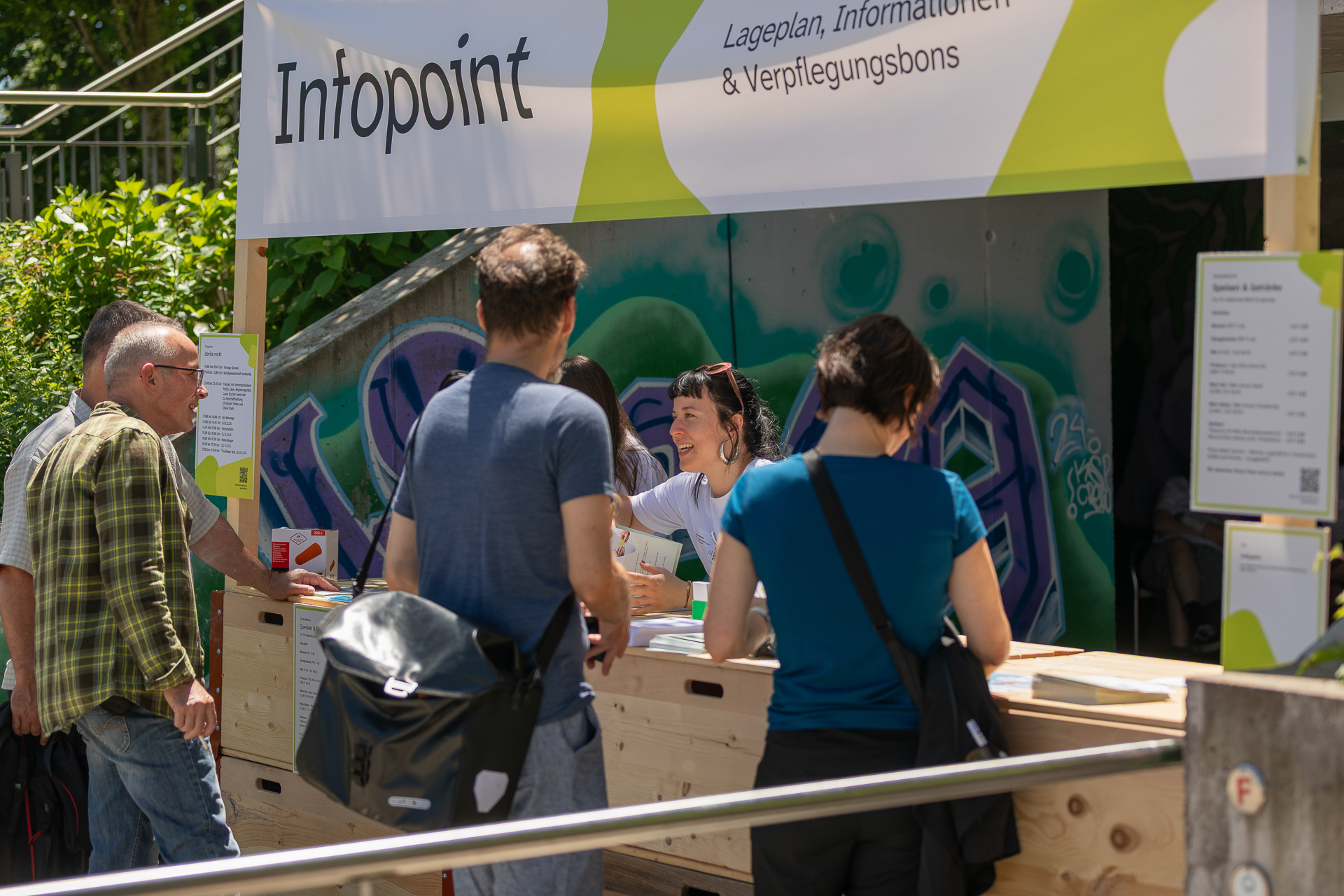 An einem Informationsstand im Freien mit der Aufschrift „Infopoint“ stehen mehrere Personen, die von einer lächelnden Frau betreut werden. Der Stand ist mit Schildern mit weiteren Informationen versehen und von Grünflächen sowie einer mit Graffiti verzierten Mauer umgeben.