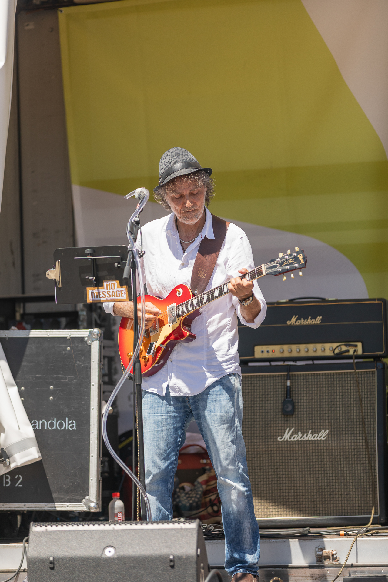 Ein Mann mit Hut und Brille spielt auf einer Außenbühne E-Gitarre und steht dabei vor Verstärkern und Tonausrüstung. Er tritt mit einem Mikrofon auf und scheint ganz in seine Musik vertieft zu sein.