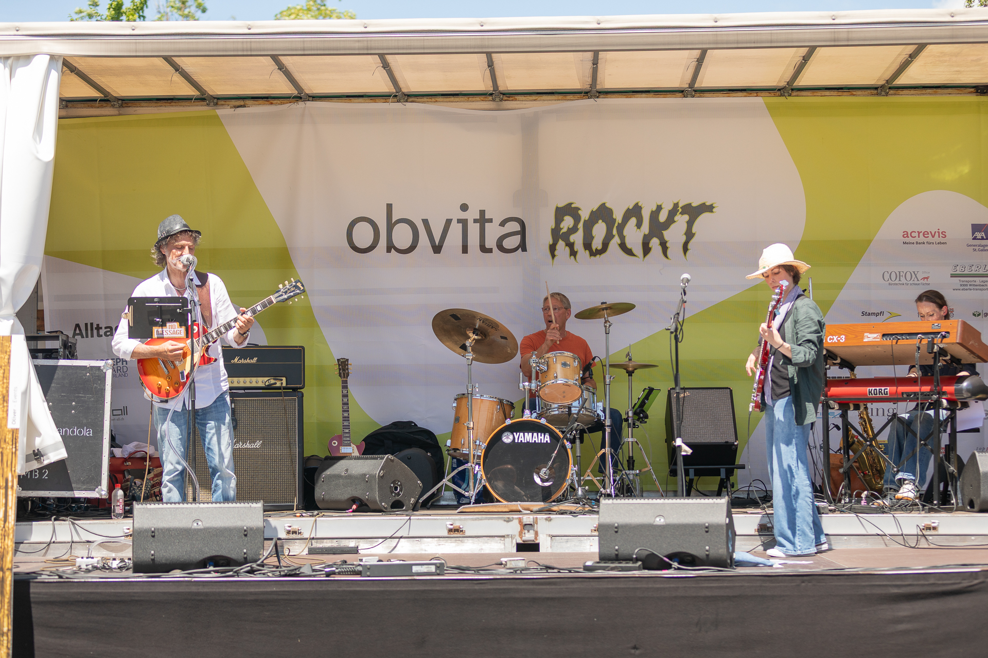 Eine Band tritt auf einer Open-Air-Bühne mit Gitarren, Schlagzeug und Keyboard auf. Auf dem Hintergrund steht „obvita ROCKT“. Die Musiker sind leger gekleidet und scheinen an einem sonnigen Tag Live-Musik zu spielen.