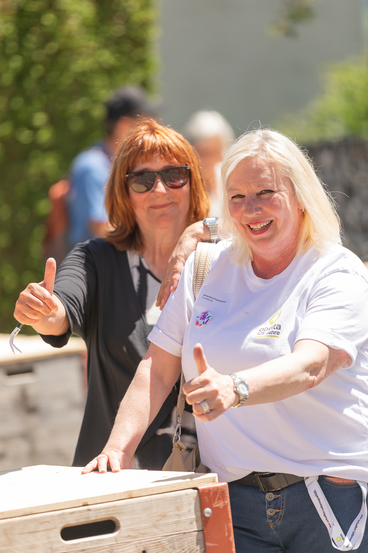 Zwei Frauen stehen im Freien, lächeln und zeigen den Daumen nach oben. Eine Frau trägt ein weißes Hemd, die andere ein dunkles Hemd und eine Sonnenbrille. Im Hintergrund sind helles Sonnenlicht und Grün zu sehen.
