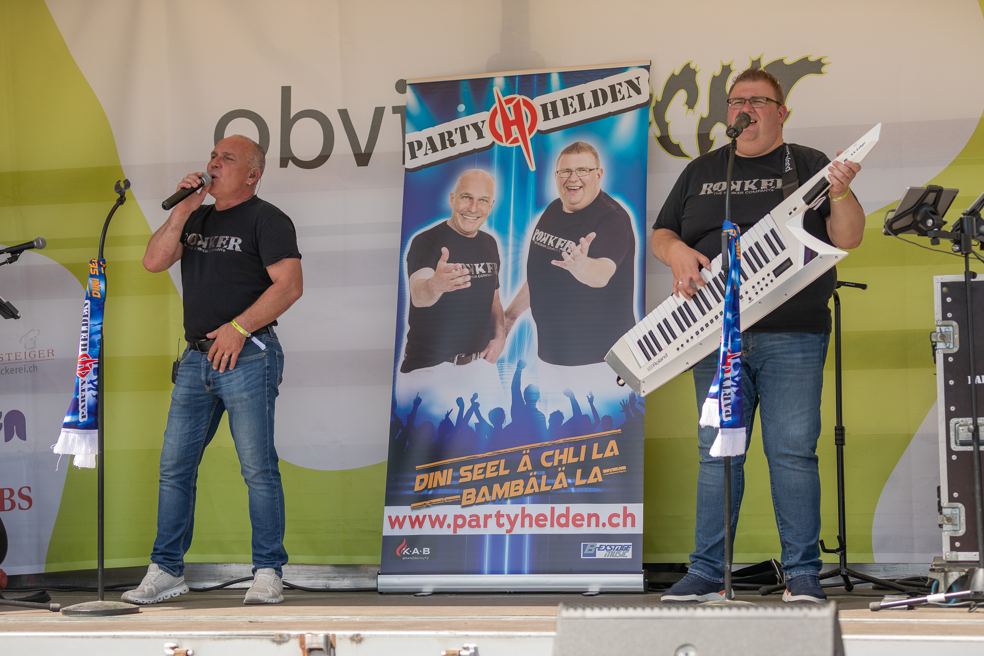 Zwei Männer treten auf der Bühne auf; der eine singt ins Mikrofon, der andere spielt Keytar. Hinter ihnen hängt ein Banner mit ihren Fotos und der Aufschrift „Party Helden“. Um sie herum sind Musikgeräte und Mikrofone zu sehen.