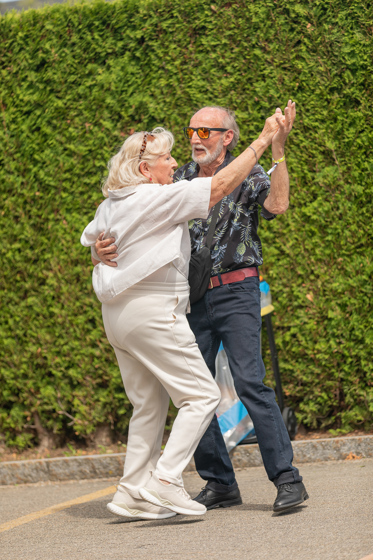 Ein älterer Mann und eine ältere Frau tanzen gemeinsam im Freien auf dem Bürgersteig, lächeln und halten sich an den Händen. Sie sind leger gekleidet, und eine grüne Hecke dient als Hintergrund.
